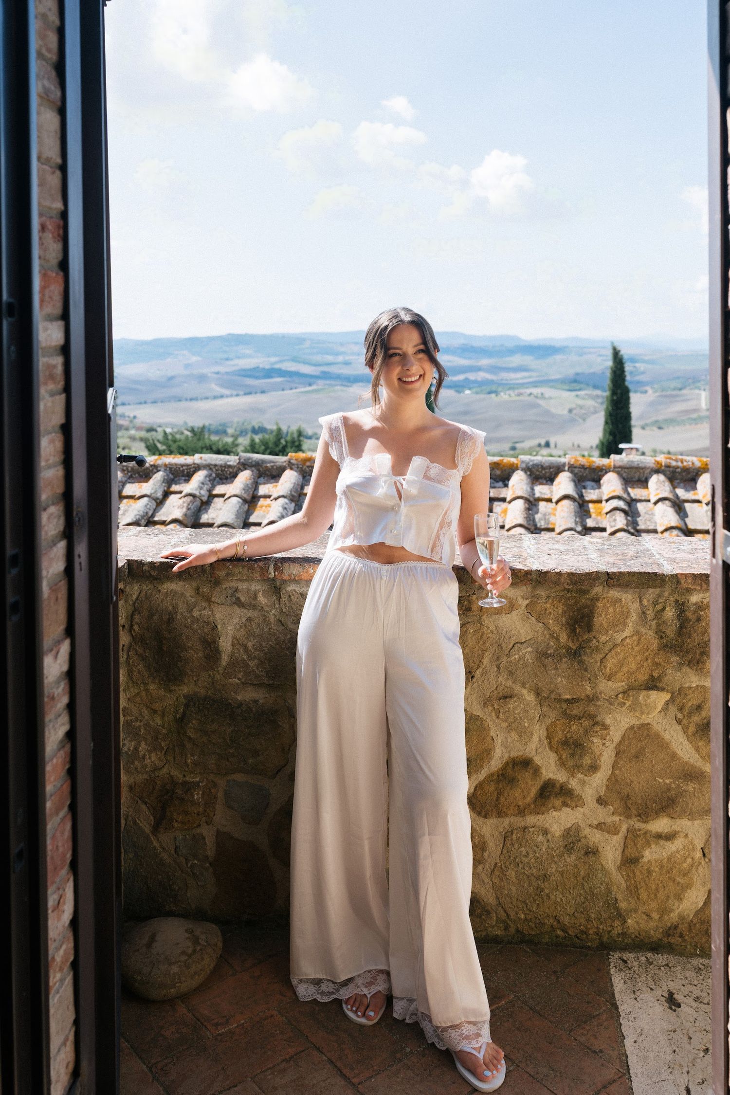Radiant bride in white crop top and wide leg trousers holding champagne on a Tuscan stone villa balcony.