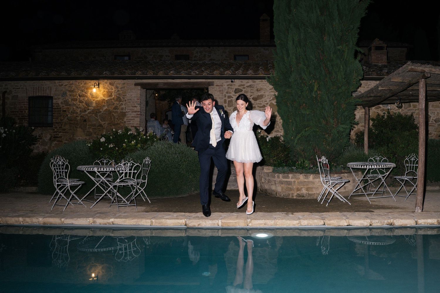 Bride and groom walking hand in hand beside illuminated pool at night.