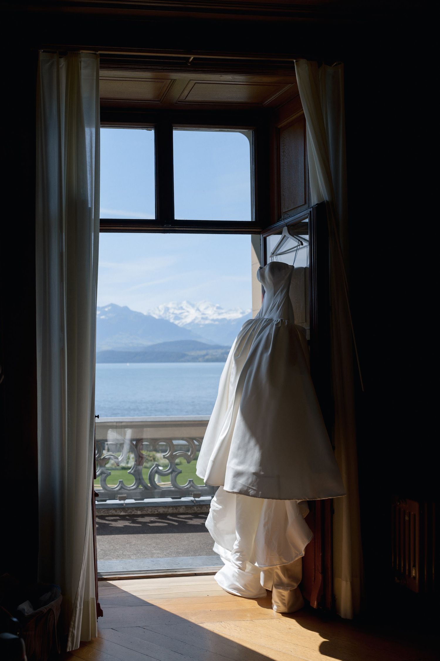White wedding dress hanging in a window with breathtaking views of a snow-capped mountain and lake.