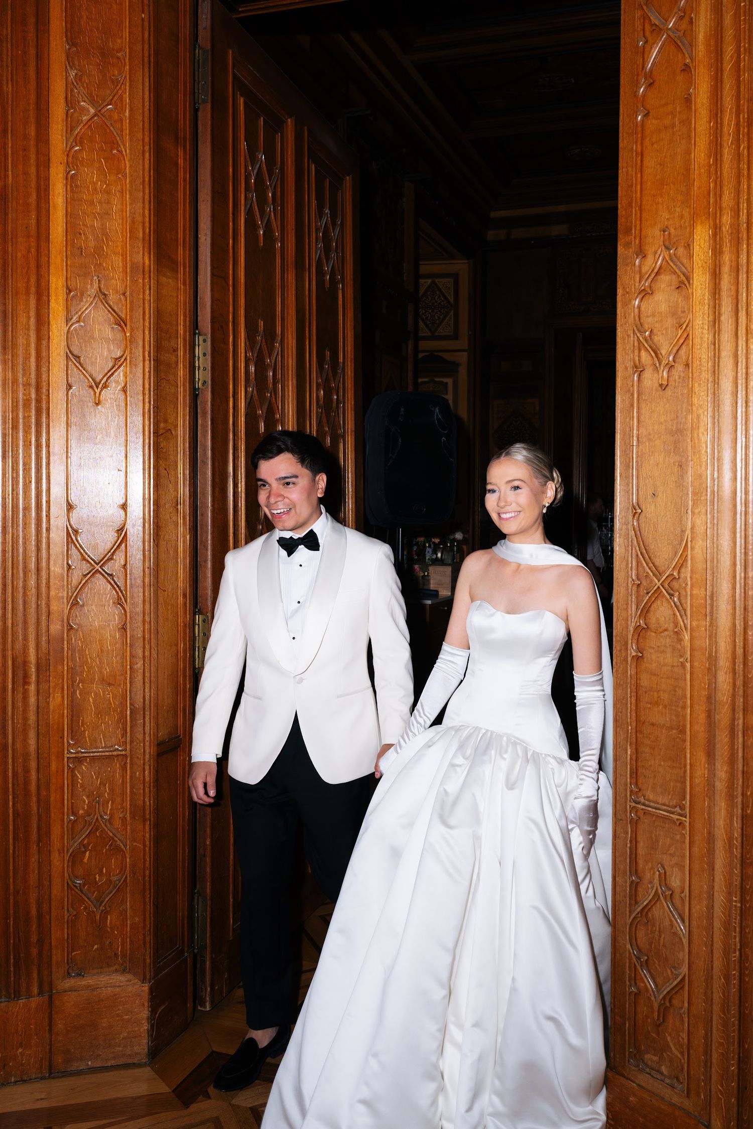 Bride in white strapless gown and groom in white tuxedo enter through ornate wooden doors smiling.