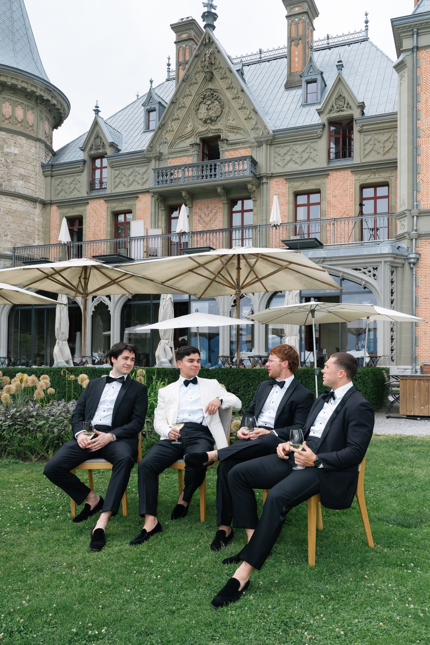 Four groomsmen in black tuxedos relaxing on benches outside an ornate château wedding venue.
