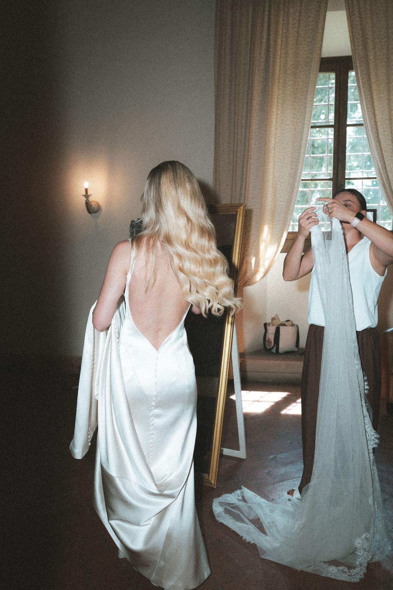 A blonde bride in a backless white satin gown stands before a gold mirror during wedding preparations.