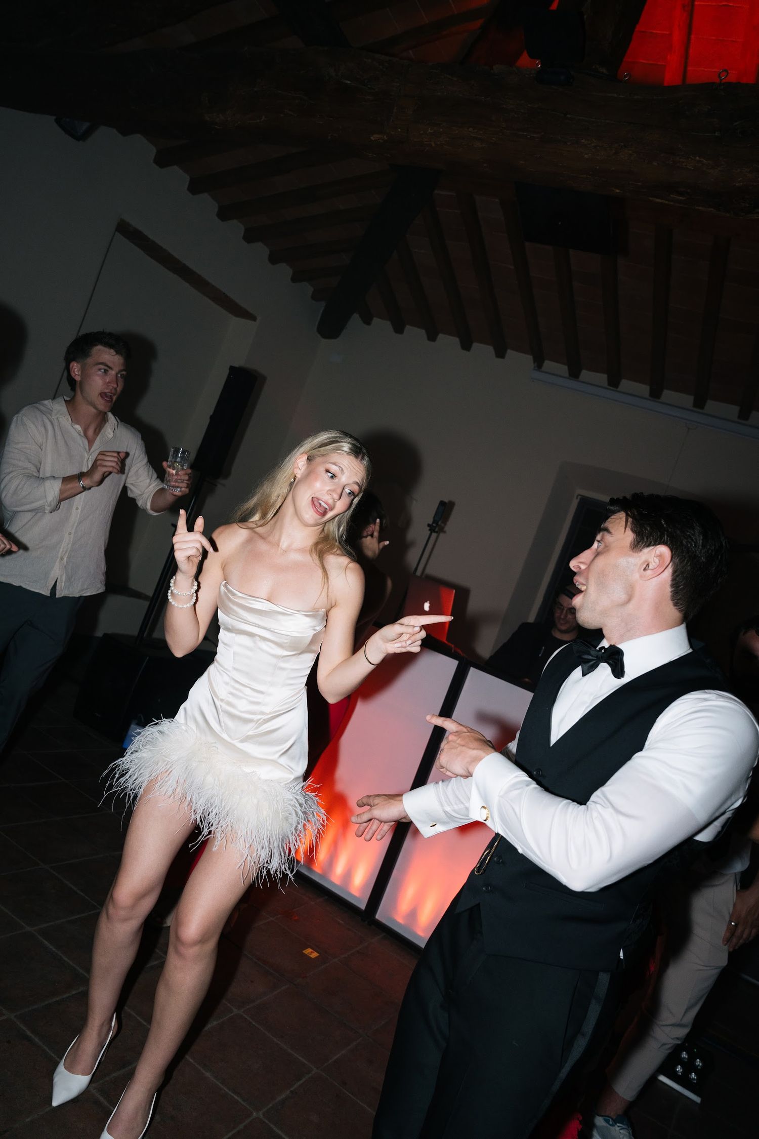 Bride in white feathered mini dress dances with groom in bow tie at wedding party.