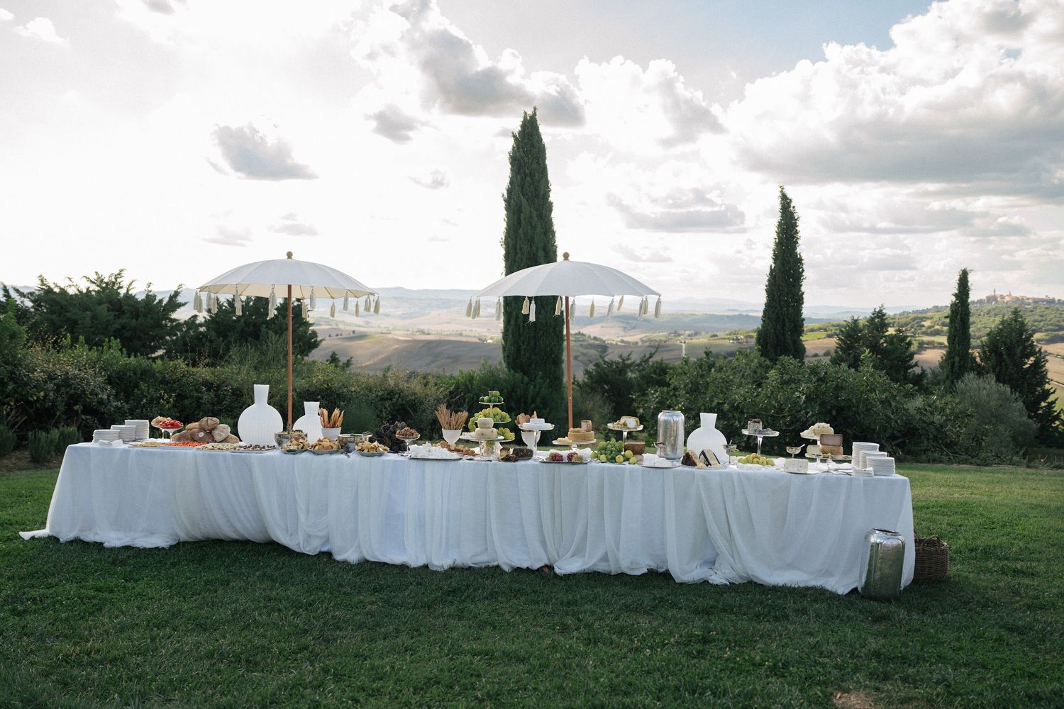 Long white draped wedding reception table with white umbrella set on green lawn with Tuscan landscape.