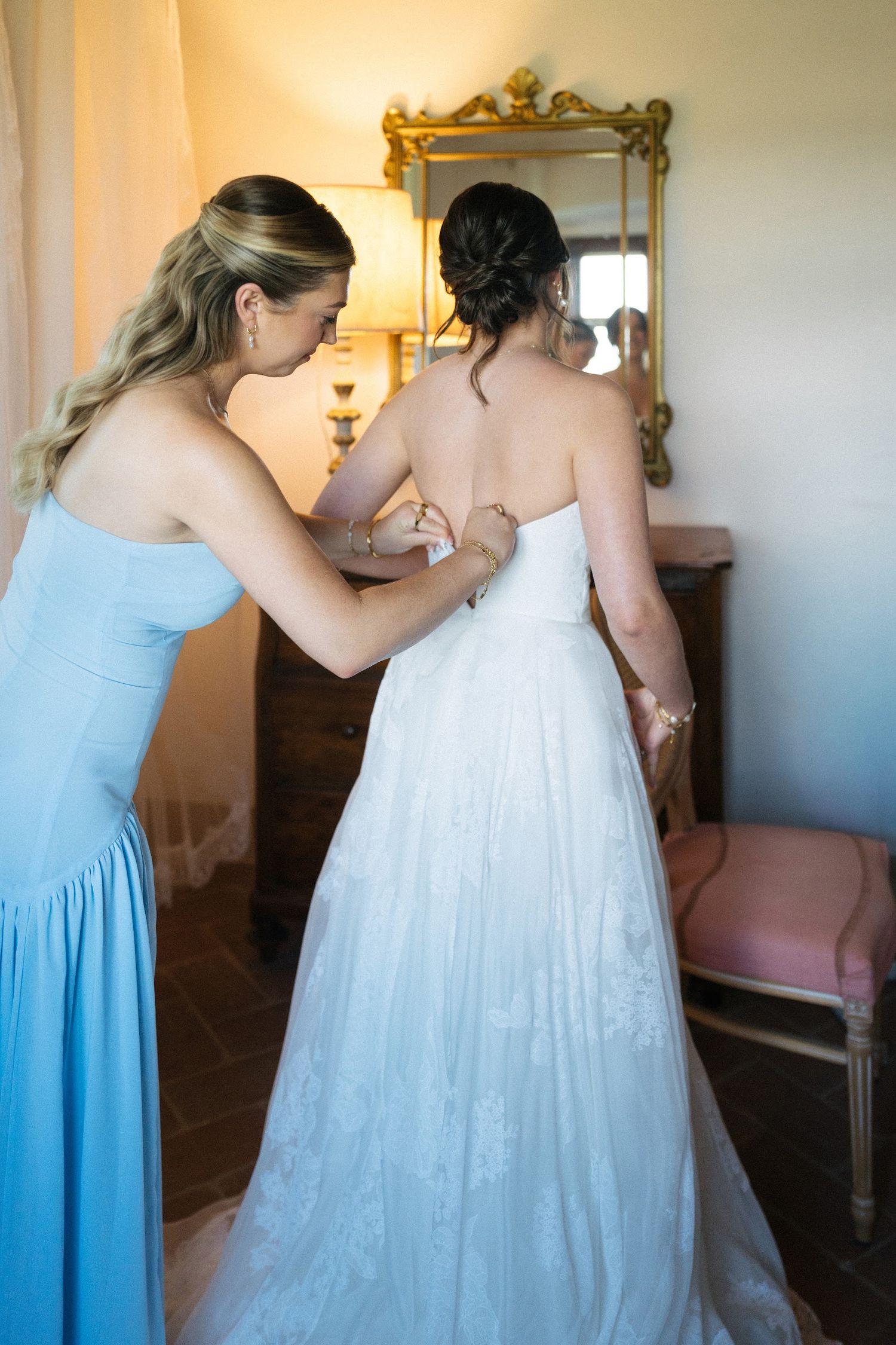 Bridesmaid in blue dress helping bride with strapless lace wedding gown in rustic Italian villa room.