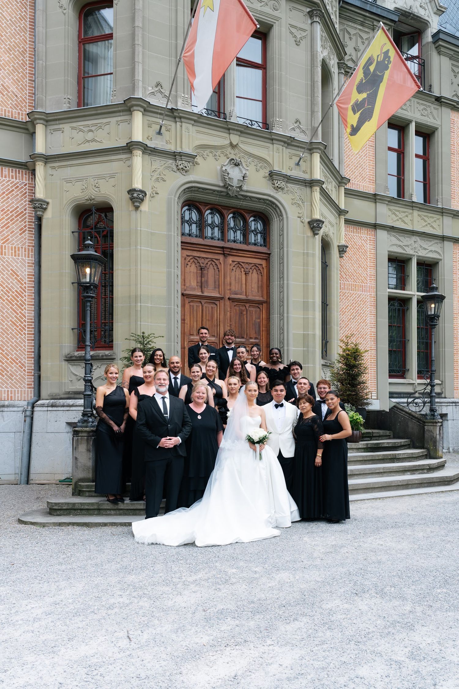 Wedding party poses before historic European building with Swiss flags, bride in white gown surrounded by guests in black.