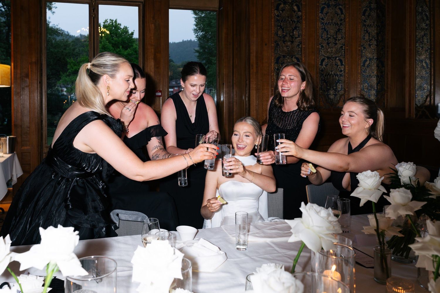 Bridesmaids in black dresses toast the bride in white at a decorated wedding reception table.