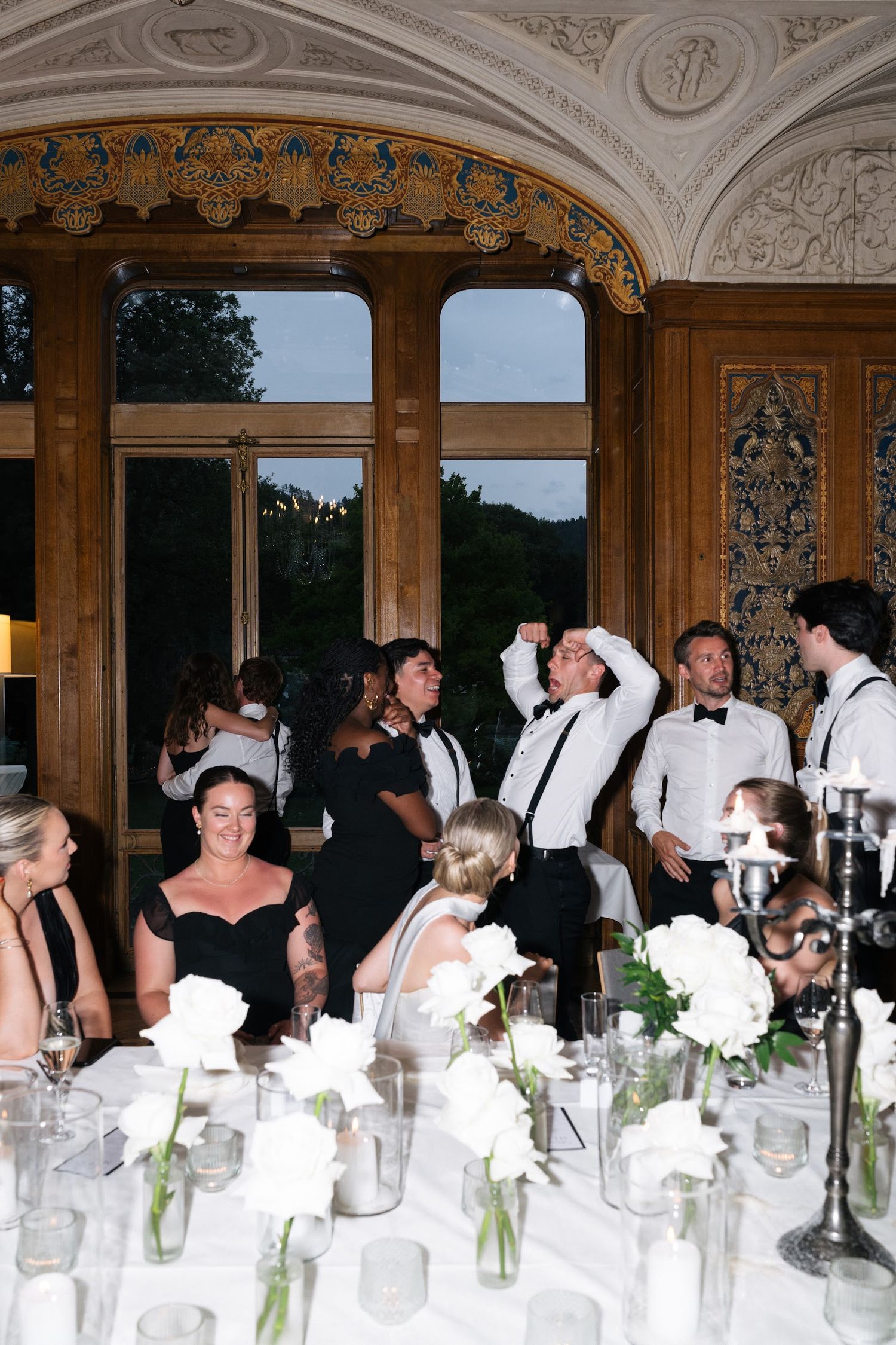 Wedding reception guests celebrating and dancing in an ornate wood-paneled ballroom with white roses.