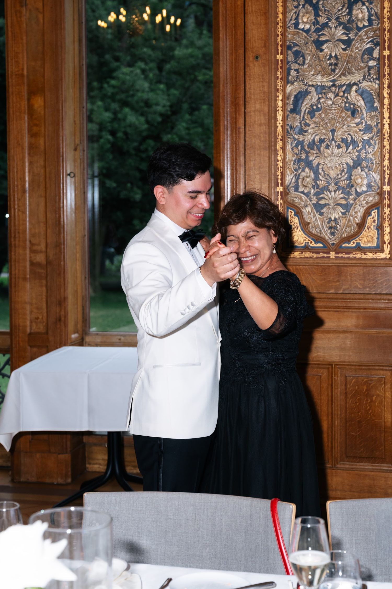 Groom in white tuxedo shares a mother-son dance in a ornate wood-paneled wedding reception venue.