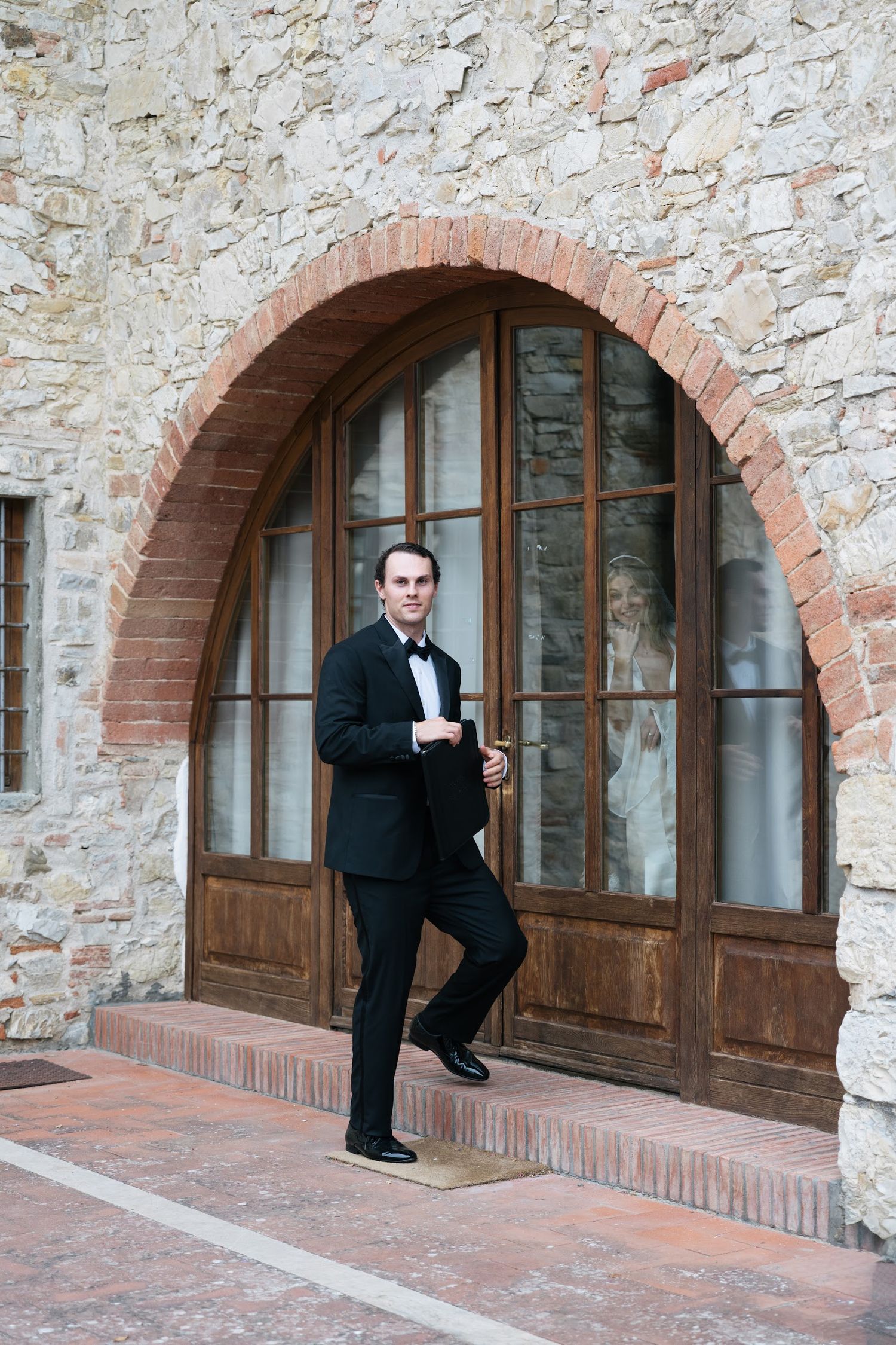 Groom in a black tuxedo stands before a rustic stone archway with wooden doors at a Tuscan venue.