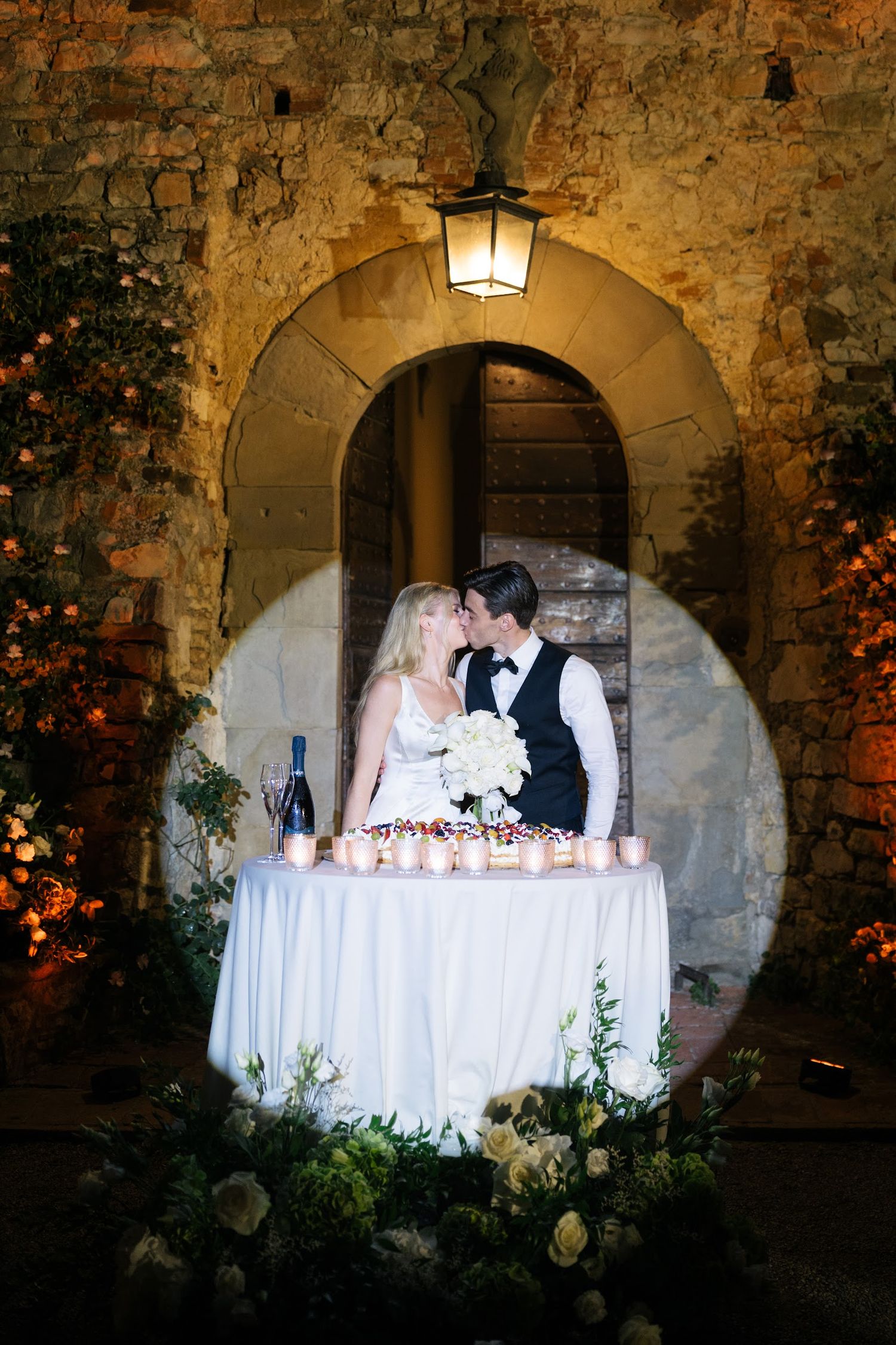 Bride and groom kissing at candlelit sweetheart table in romantic Italian castle archway.