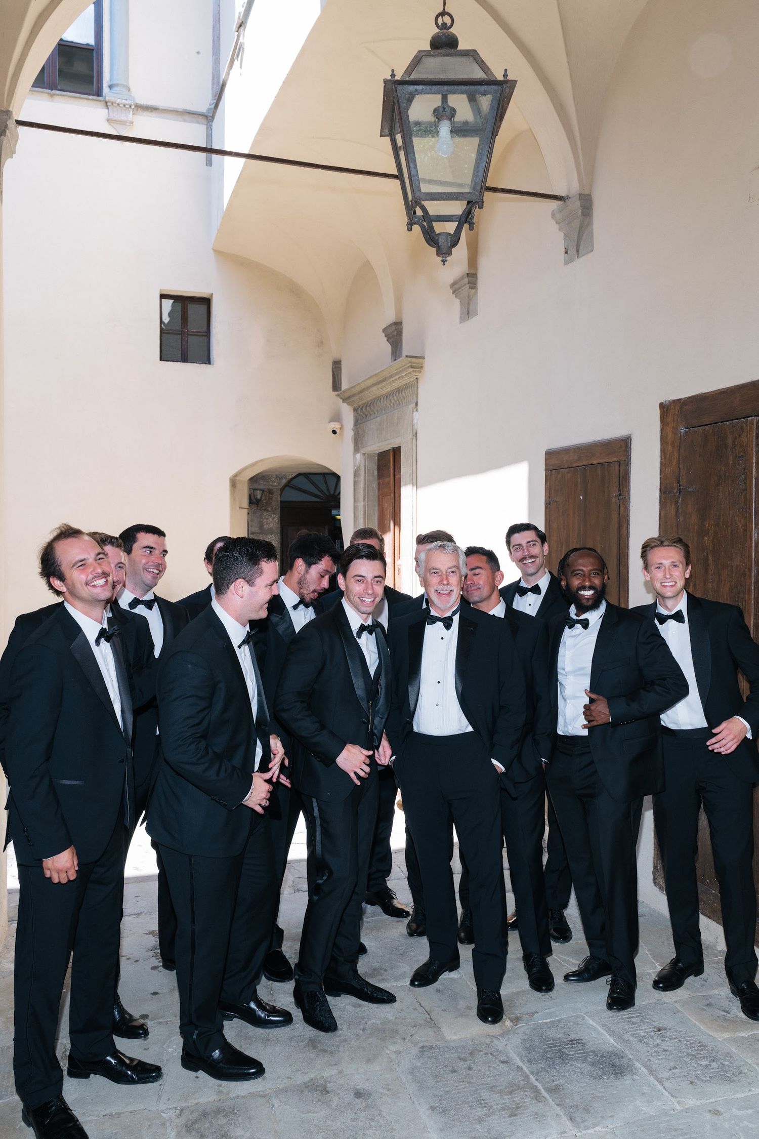 Groom and groomsmen in black tuxedos laughing together in a sunlit courtyard of an Italian castle.