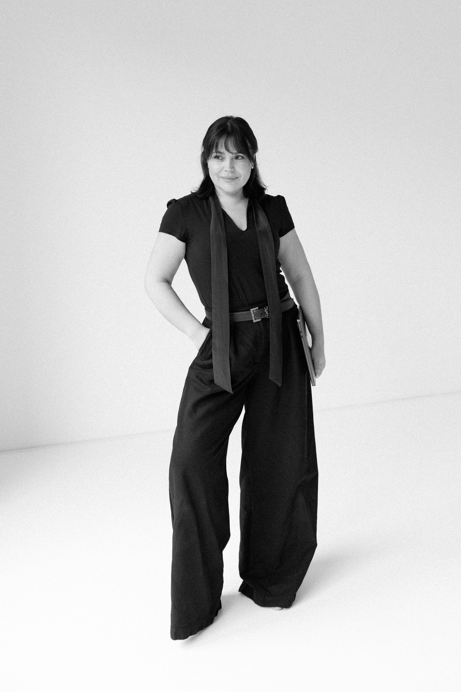 Woman in black wide-leg trousers and top poses confidently in minimalist black and white studio portrait.