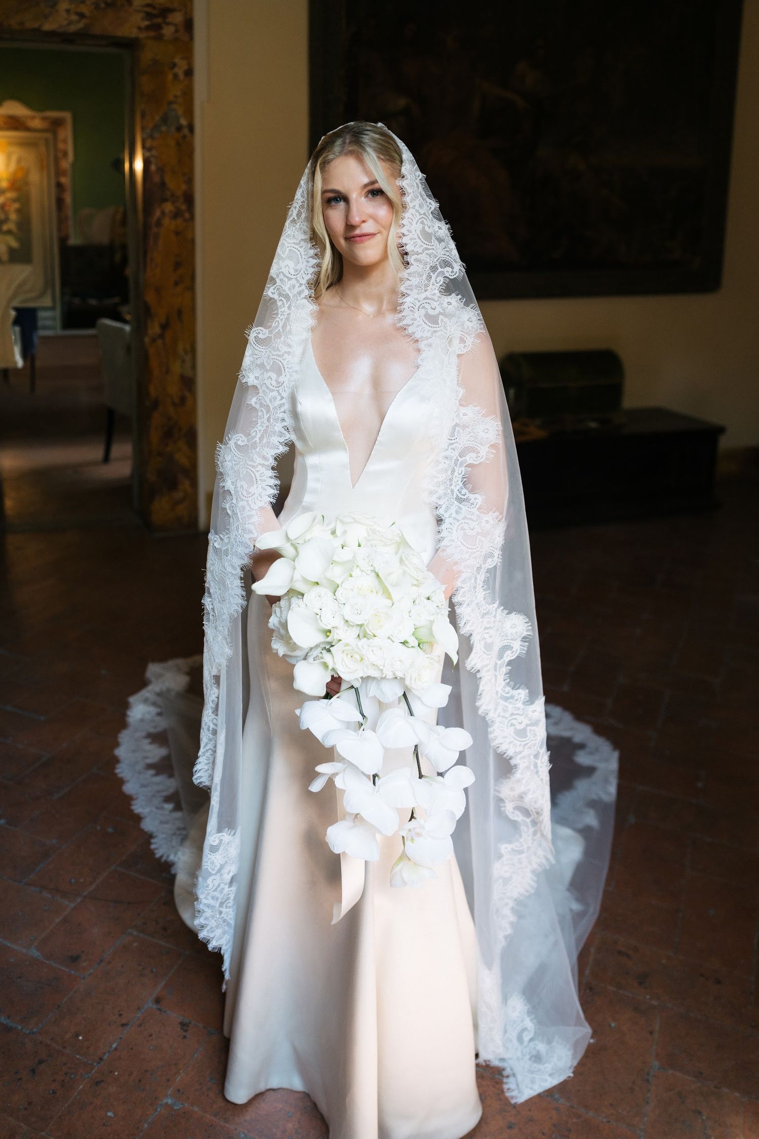 Bride in deep V-neck gown with lace-trimmed cathedral veil holding cascading white orchid bouquet indoors.