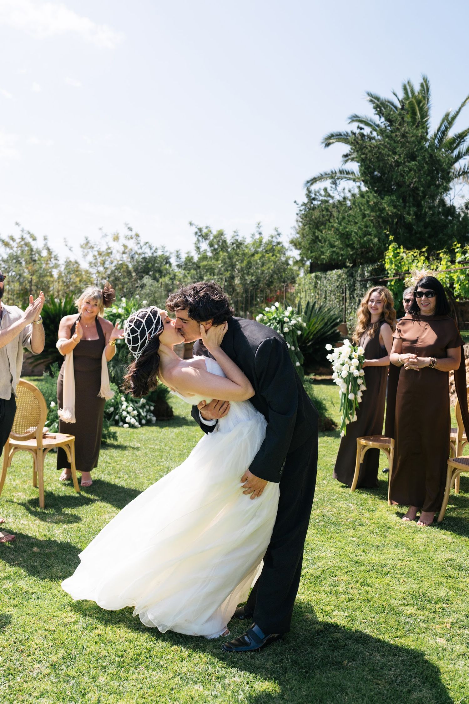 Groom dips bride for a kiss at outdoor garden wedding ceremony while guests applaud.