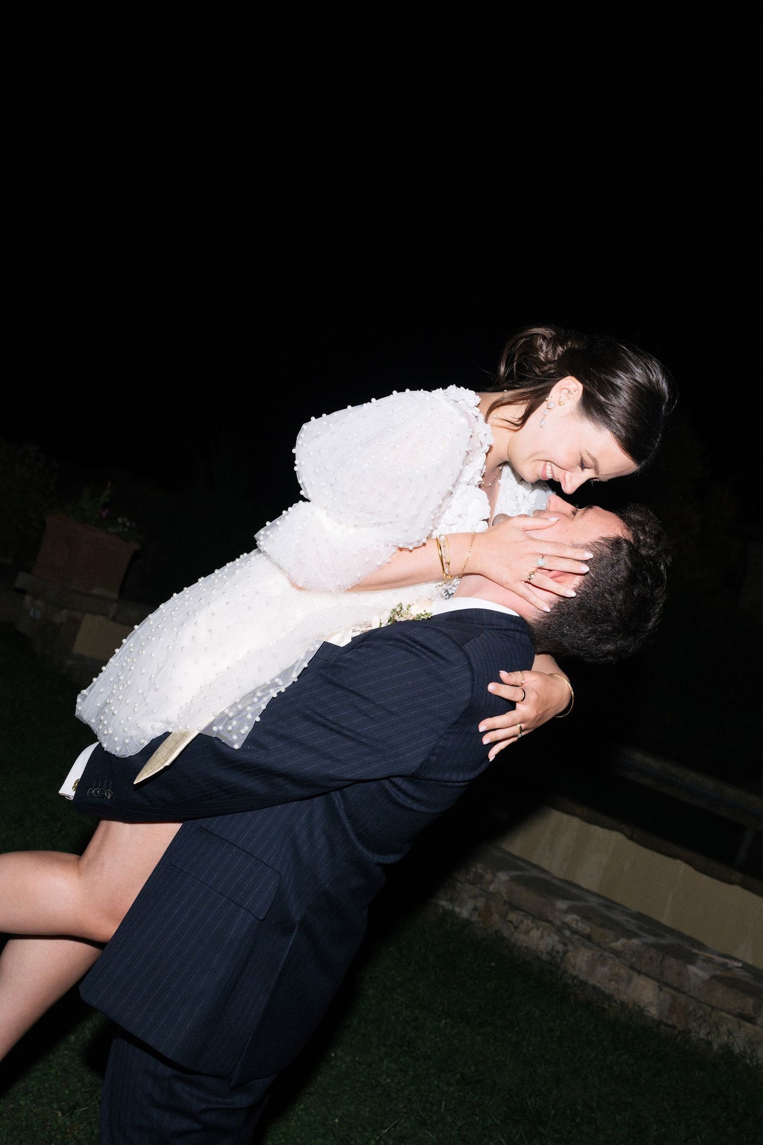 Groom lifting bride in white pearl dress outdoors at night wedding.