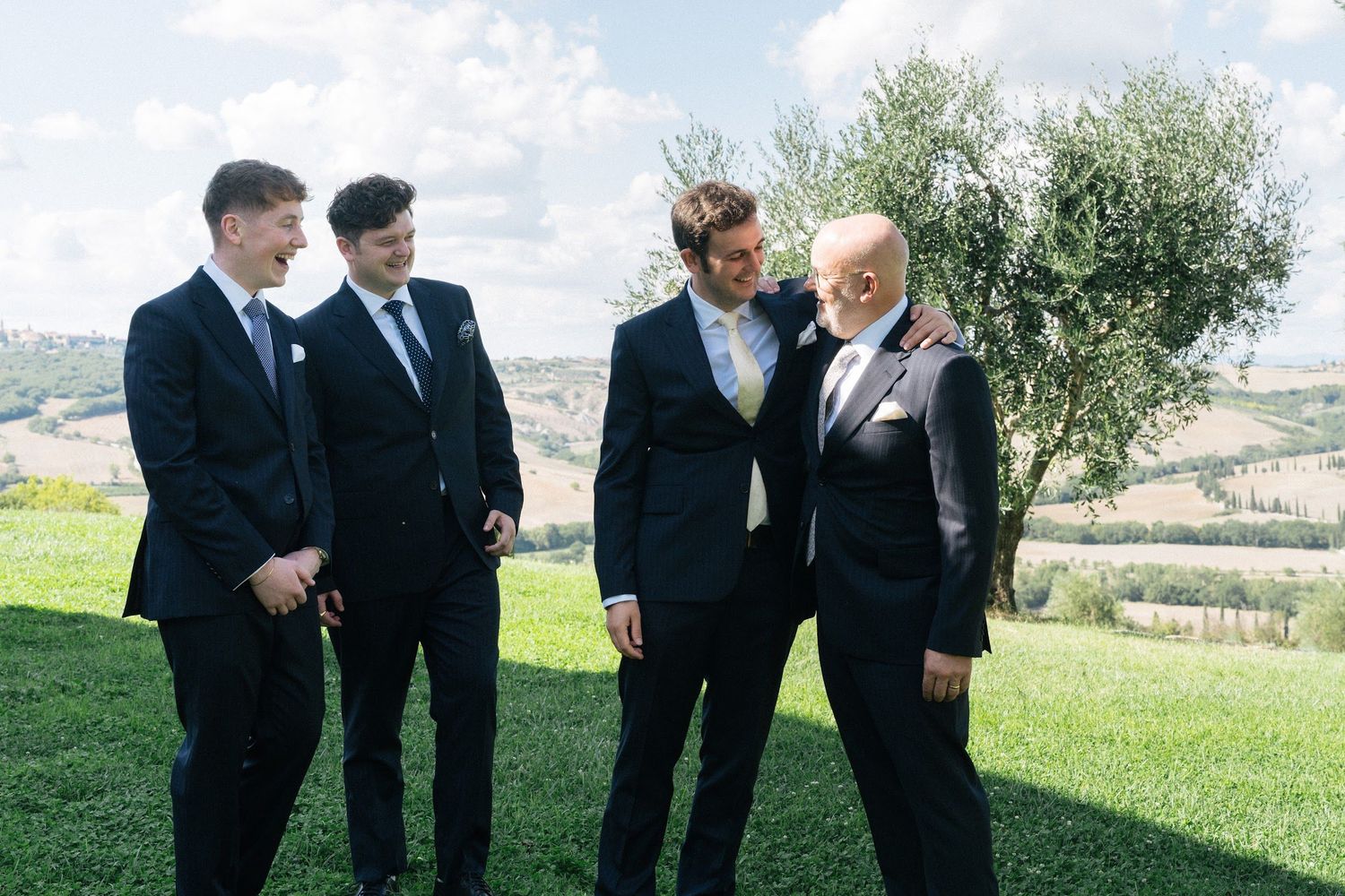 Groom and three groomsmen in dark suits laughing together on a green hillside in the Tuscan countryside.
