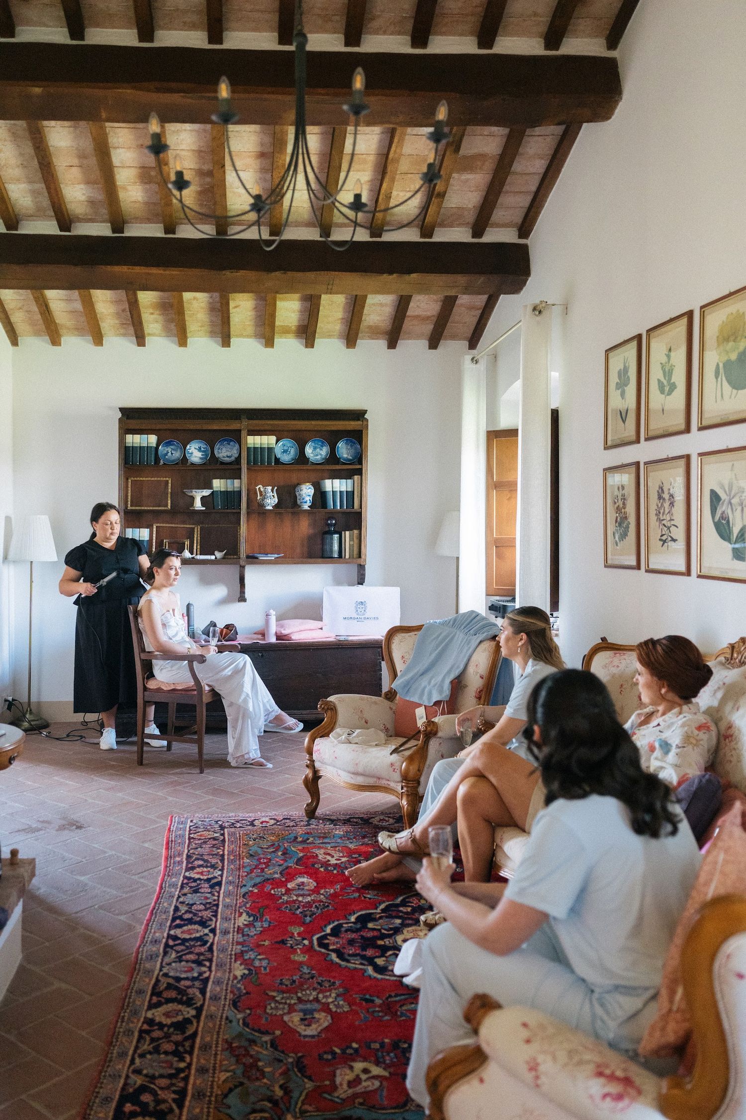 Bride getting ready with bridesmaids in a rustic Tuscan villa interior with wooden beamed ceiling and antique furniture.