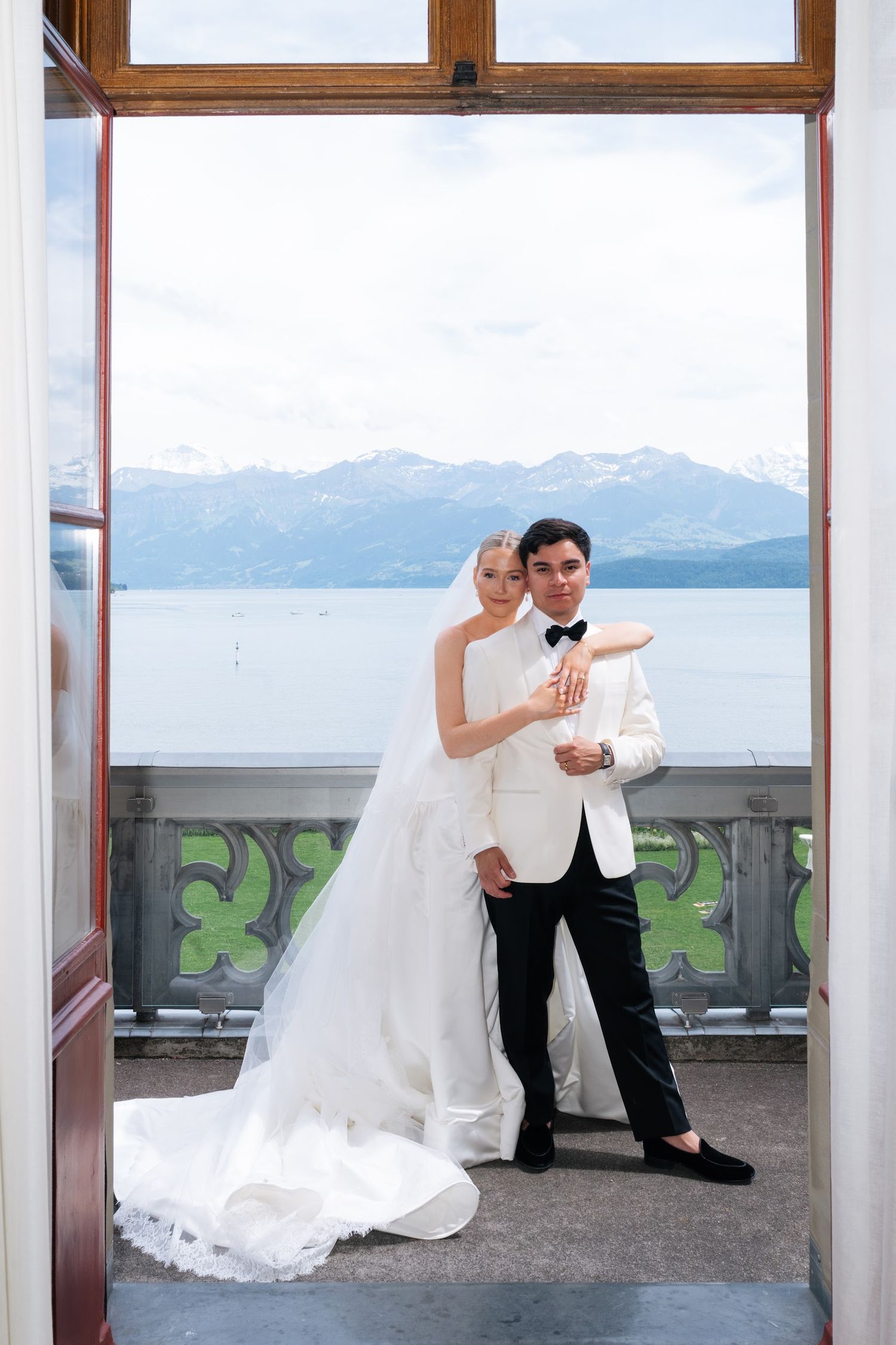 Bride and groom pose in open doorway with panoramic alpine lake and mountain views, groom in white dinner jacket.