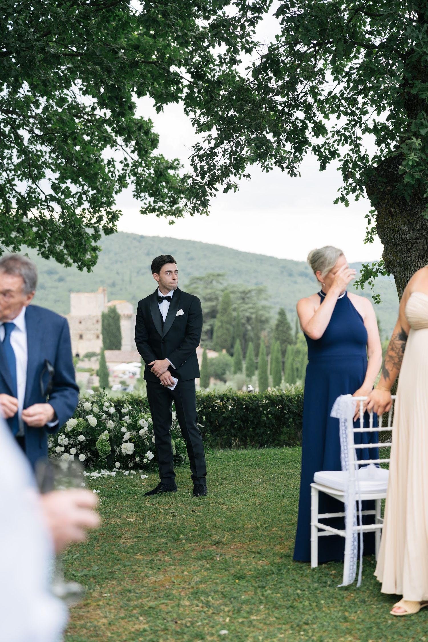Groom in black tuxedo stands at outdoor wedding altar as emotional guest wipes tears, Tuscan hills in background.