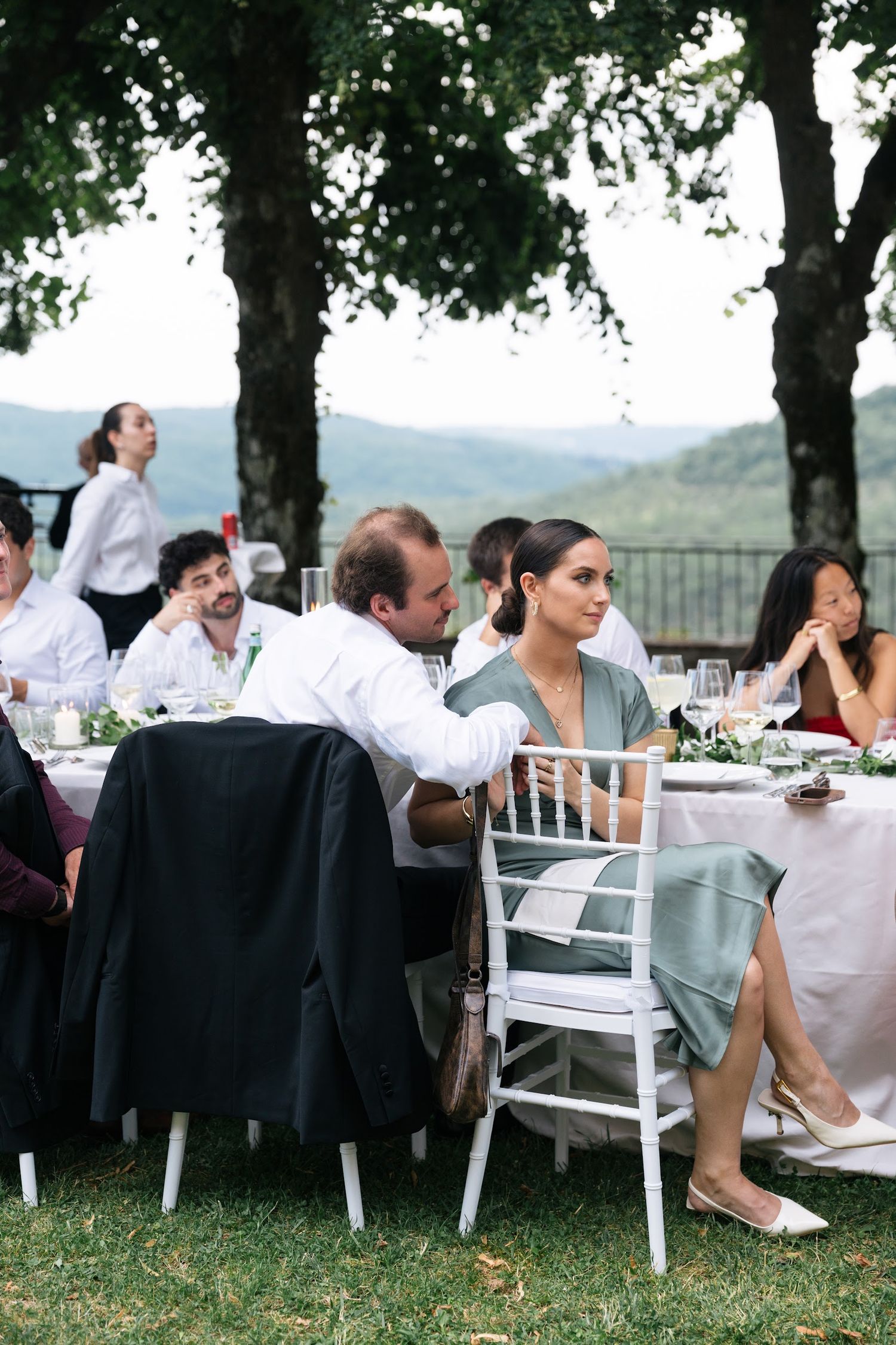 Wedding guests seated at outdoor reception tables with scenic hillside views in background.