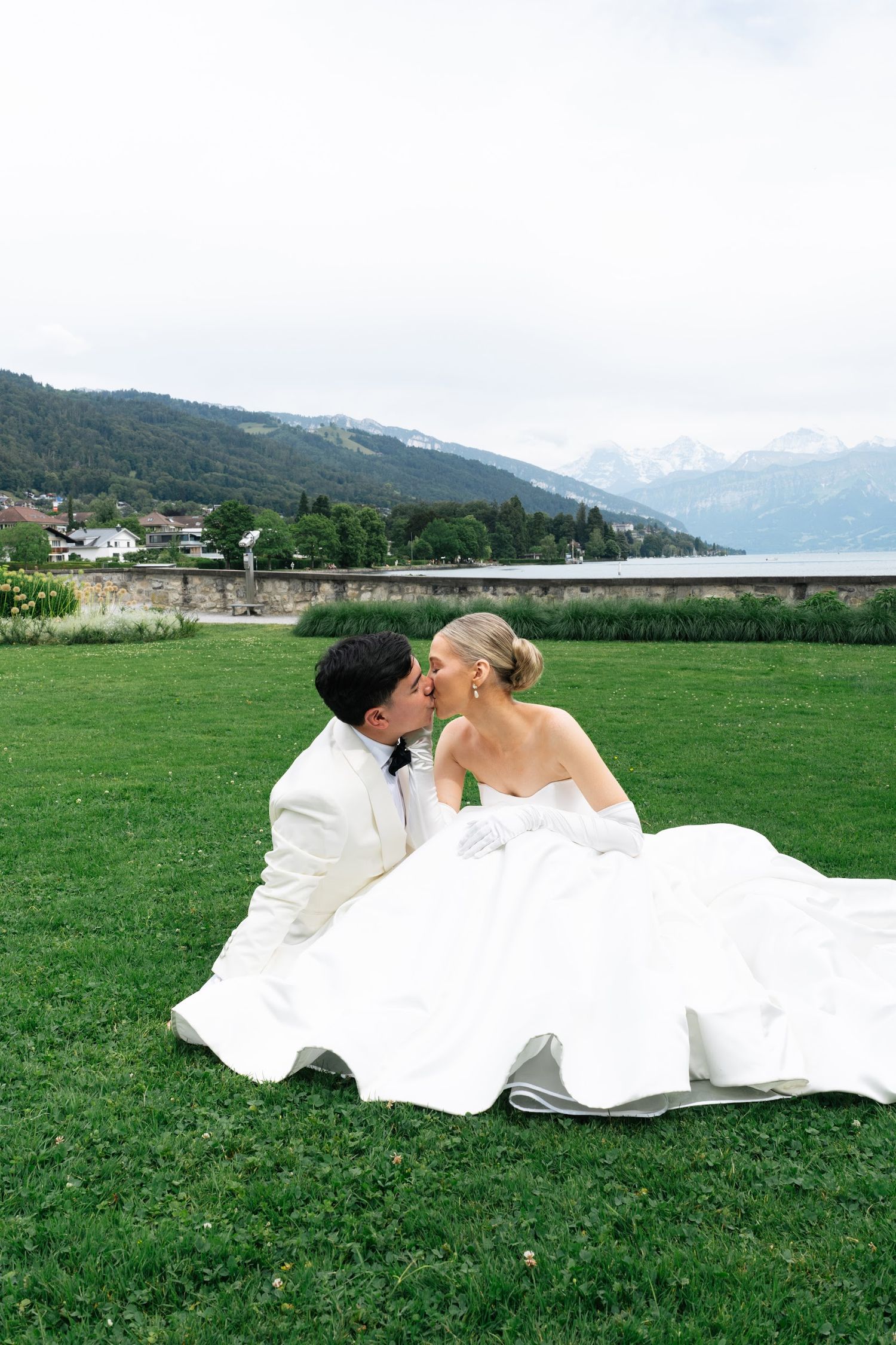 Bride and groom sharing a kiss while sitting on green lawn with scenic mountain lake behind them.