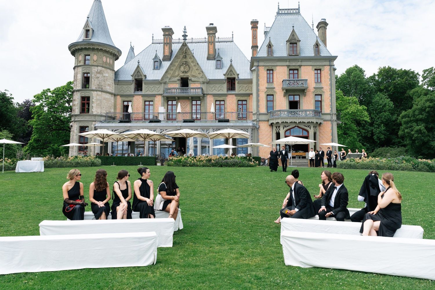 Guests in black attire seated on white couches on a lawn before a grand château during an outdoor wedding reception.
