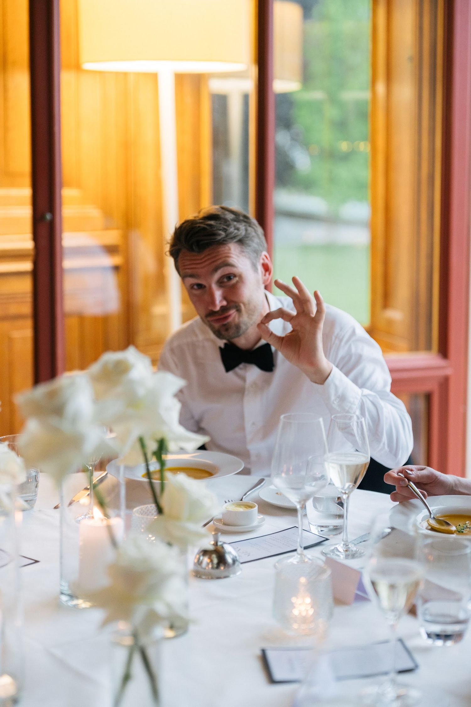 Dapper wedding guest in bow tie gives an OK hand gesture at elegant reception dinner table.