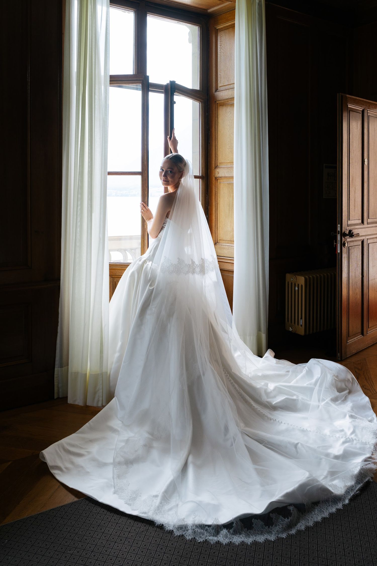 Bride in stunning white ball gown with long lace-trimmed veil gazing out tall window in elegant wood-paneled room.