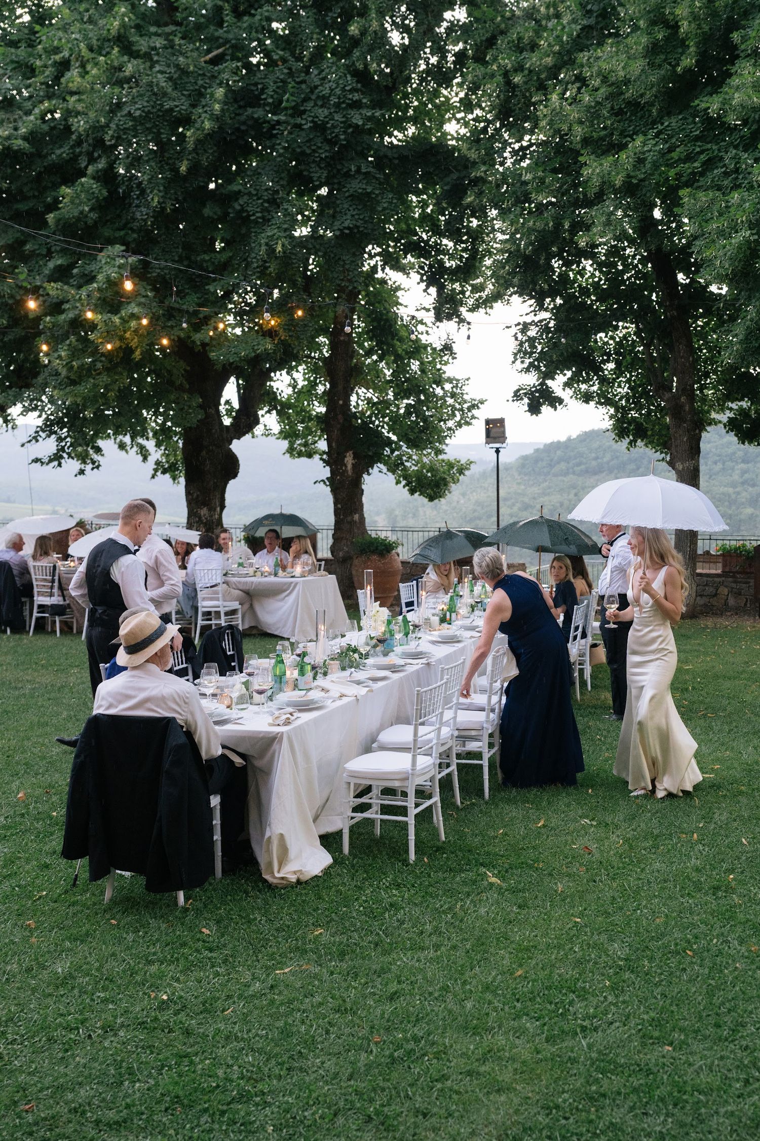Wedding guests holding white umbrellas at elegant outdoor reception as light rain falls.