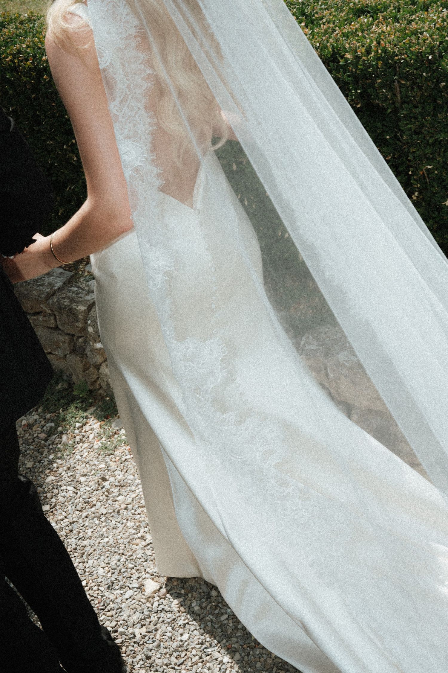 Close-up of a bride in a fitted lace wedding gown with a long flowing cathedral veil outdoors.