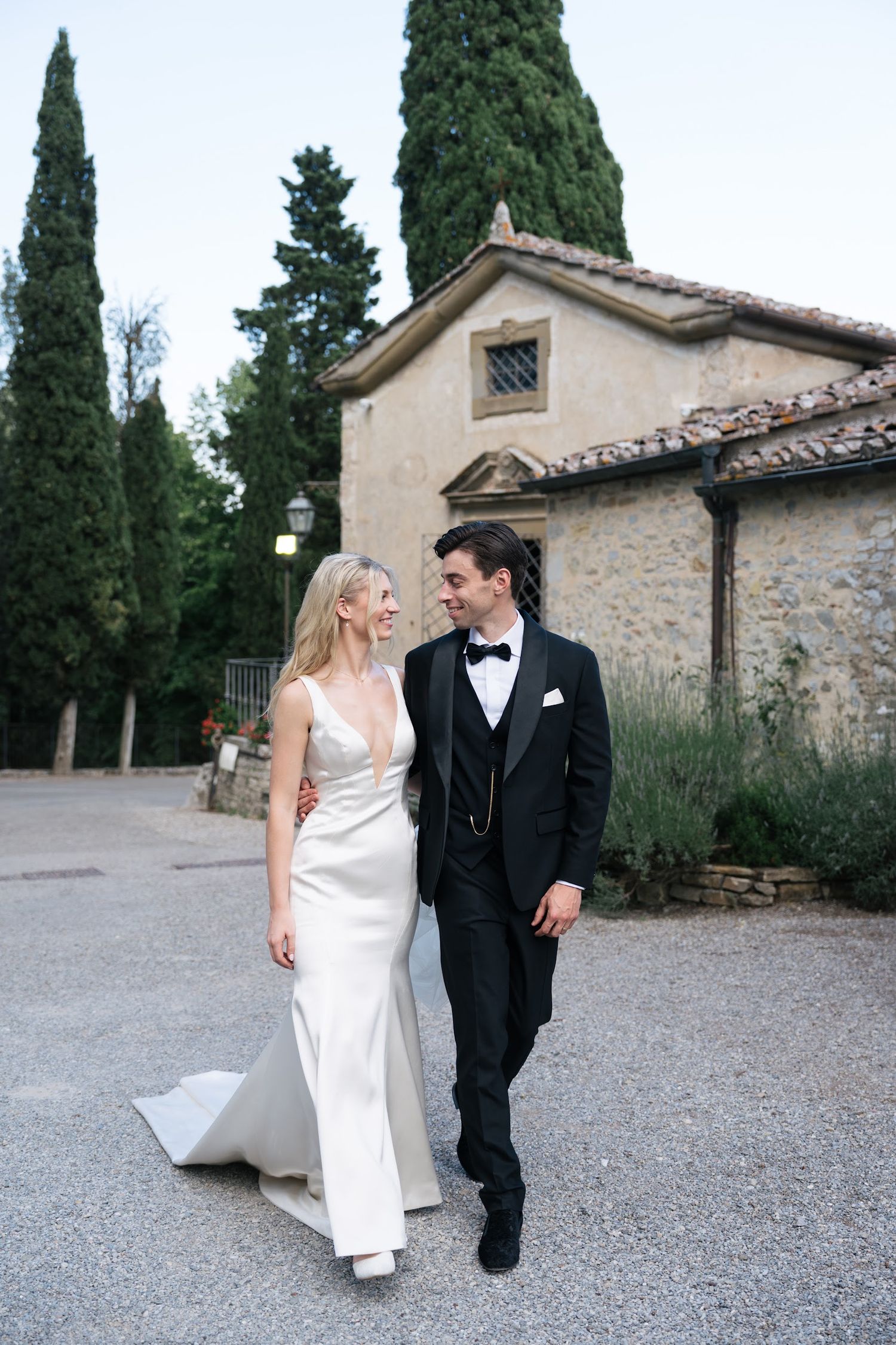 Bride in sleek white gown and groom in black tuxedo walk and smile outside a rustic Tuscan stone chapel.