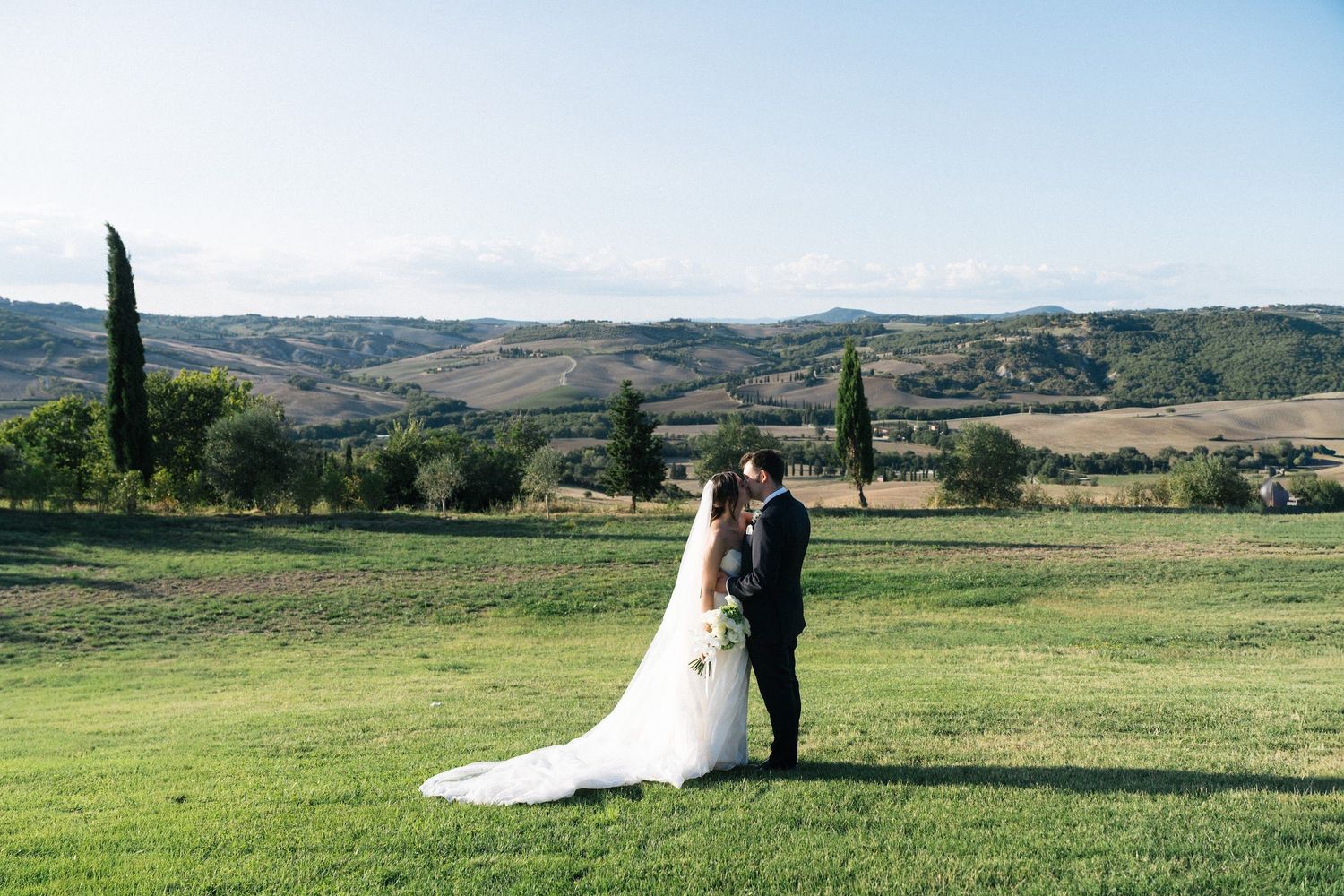 Bride and groom share romantic moment on lush green lawn with rolling Tuscan hills behind them.