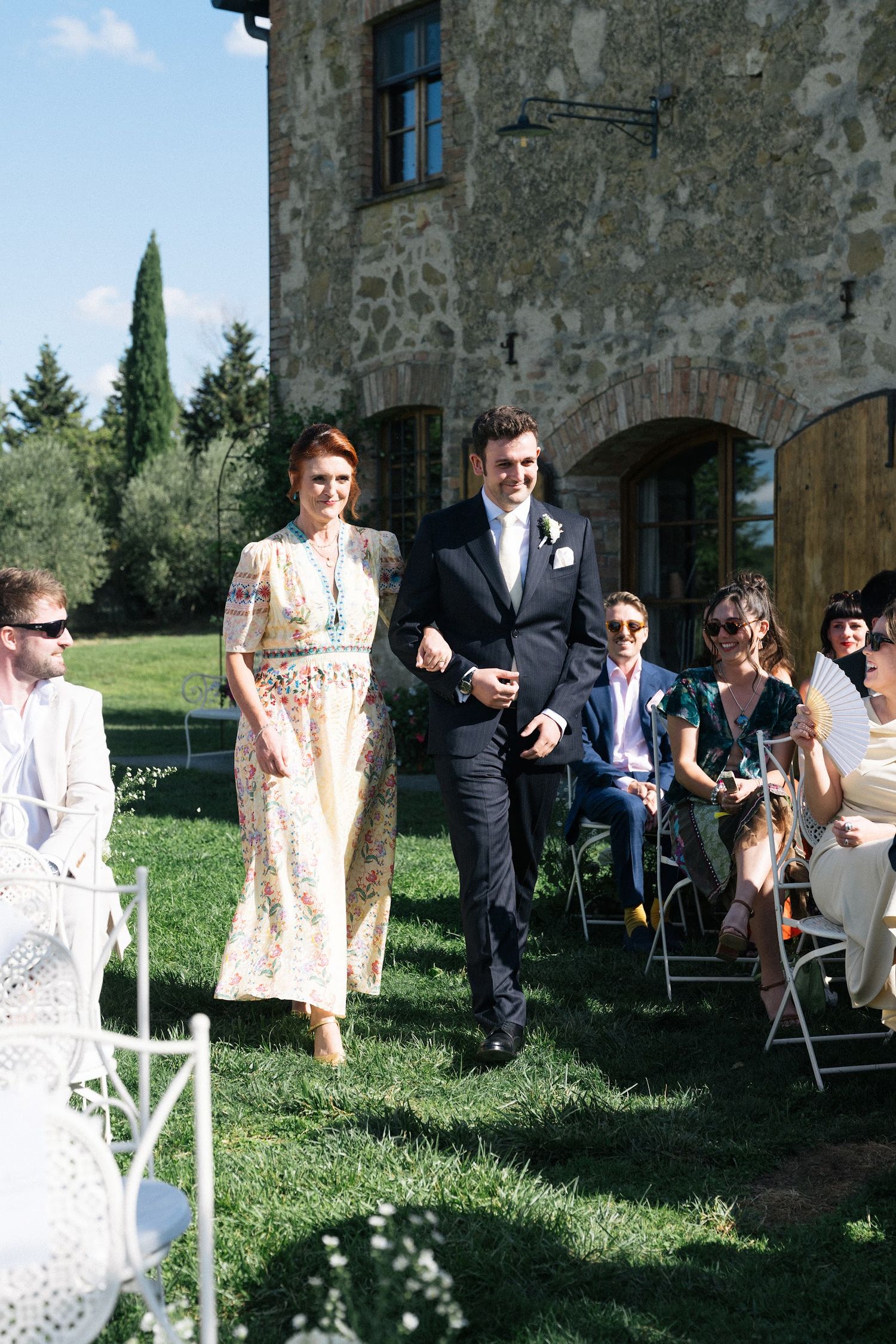 Groom in dark suit walking down outdoor aisle with woman in floral dress at Tuscan stone villa wedding.