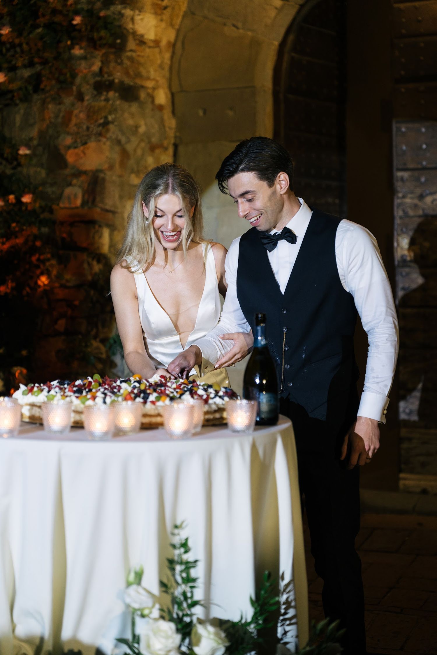 Bride and groom smiling while cutting their wedding cake at an elegant candlelit reception table.