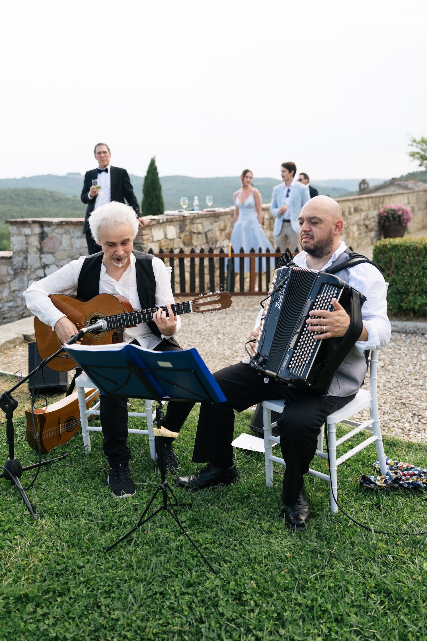 Two musicians play guitar and accordion at an outdoor Tuscan wedding reception with rolling hills behind them.