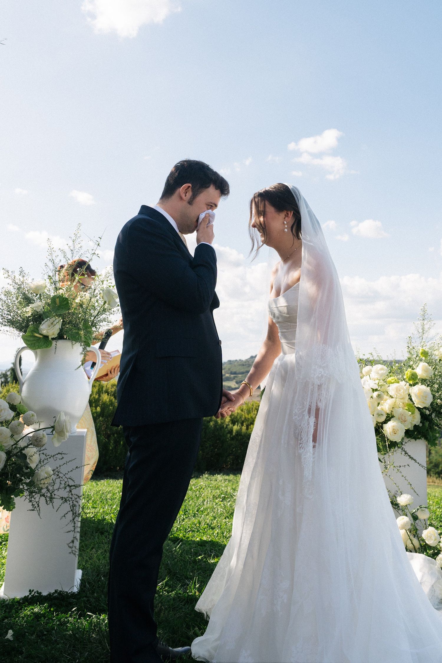 Emotional groom wiping tears holding brides hands during outdoor wedding ceremony with white floral arrangements.