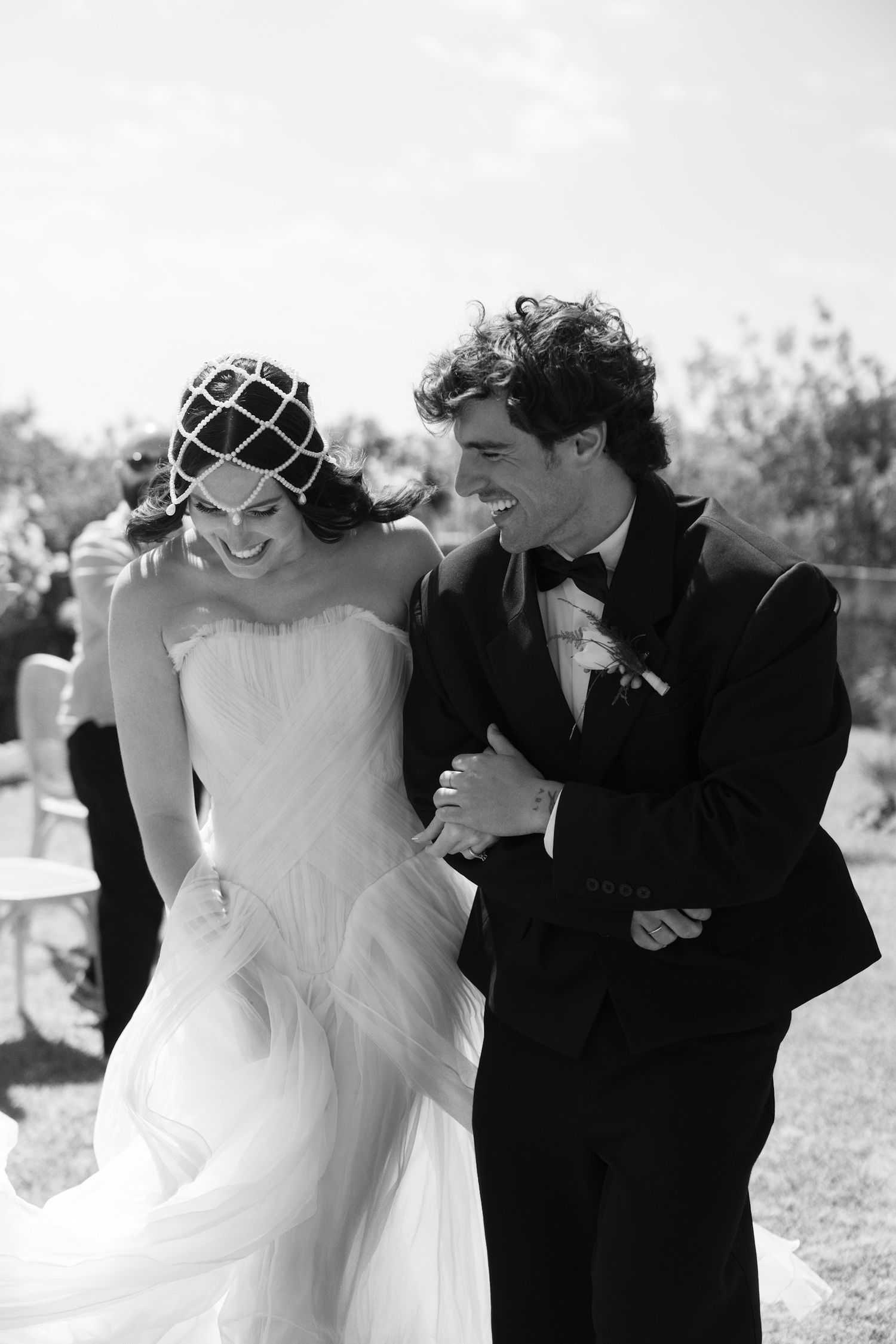 Black and white photo of laughing bride and groom in tuxedo walking arm in arm outdoors.