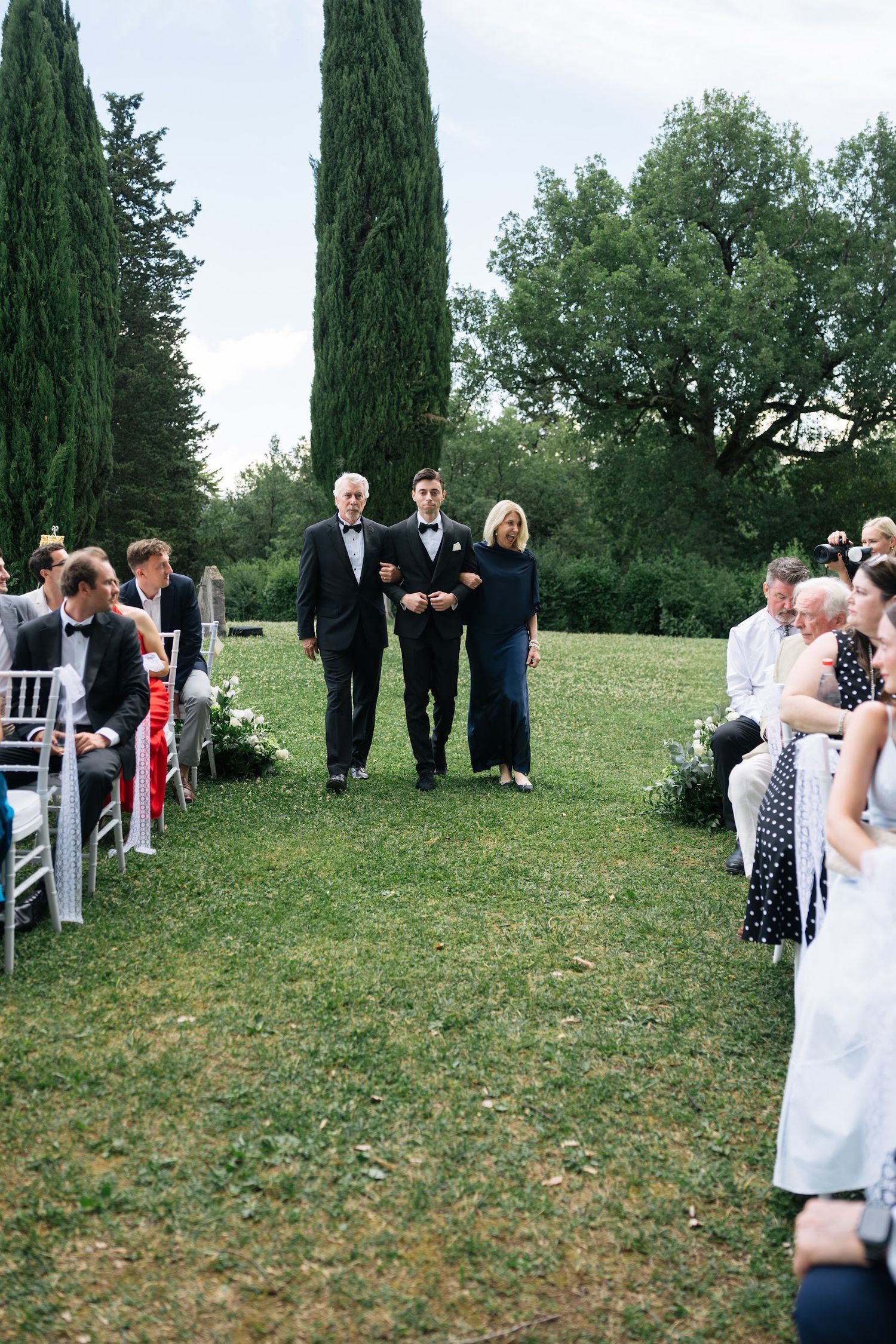 Groom escorted down grassy aisle by parents during elegant outdoor wedding ceremony in Tuscany, Italy.