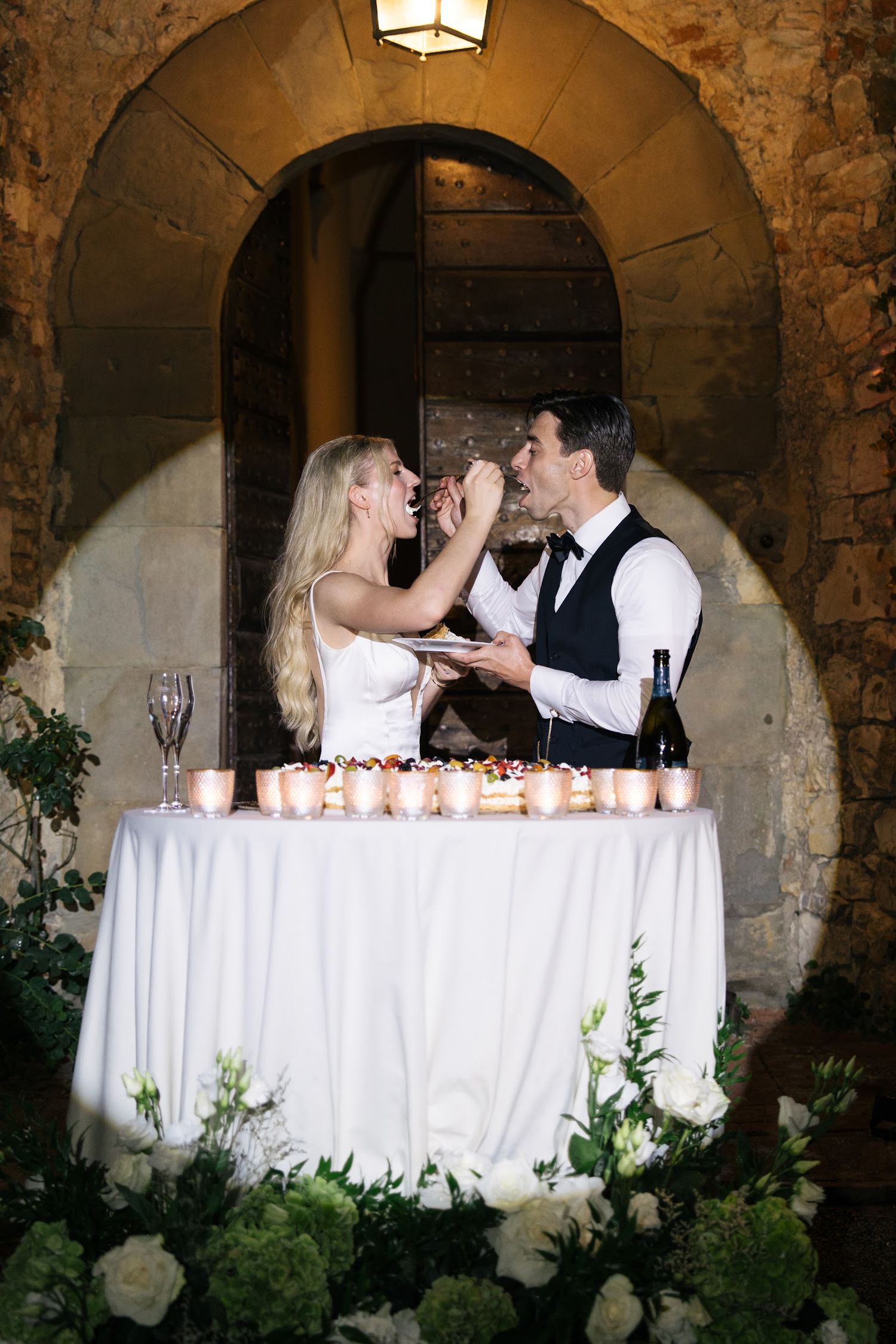 Bride feeding groom wedding cake at a candlelit sweetheart table adorned with white floral arrangements.