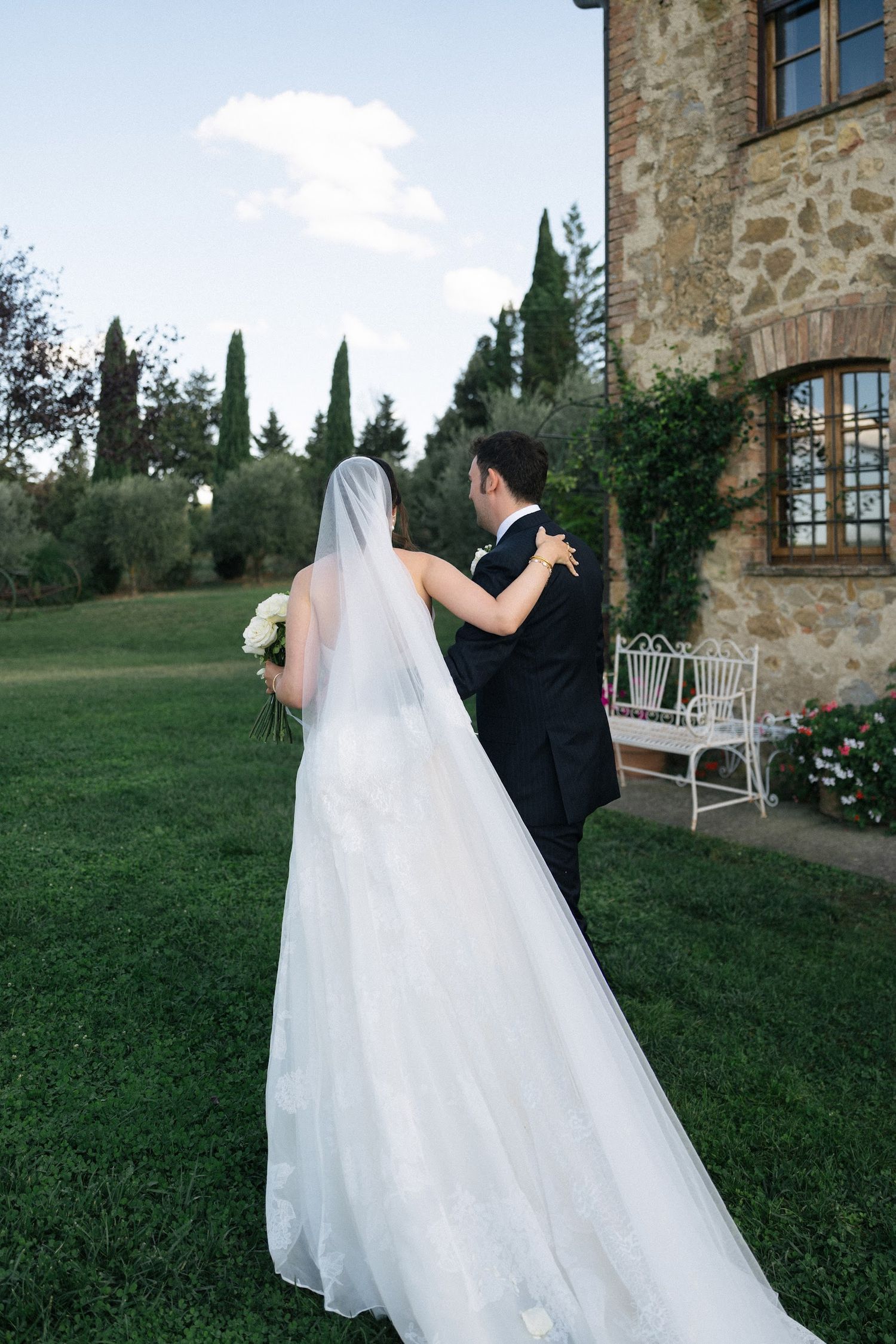 Bride and groom walking hand in hand toward rustic stone villa with long cathedral veil trailing.
