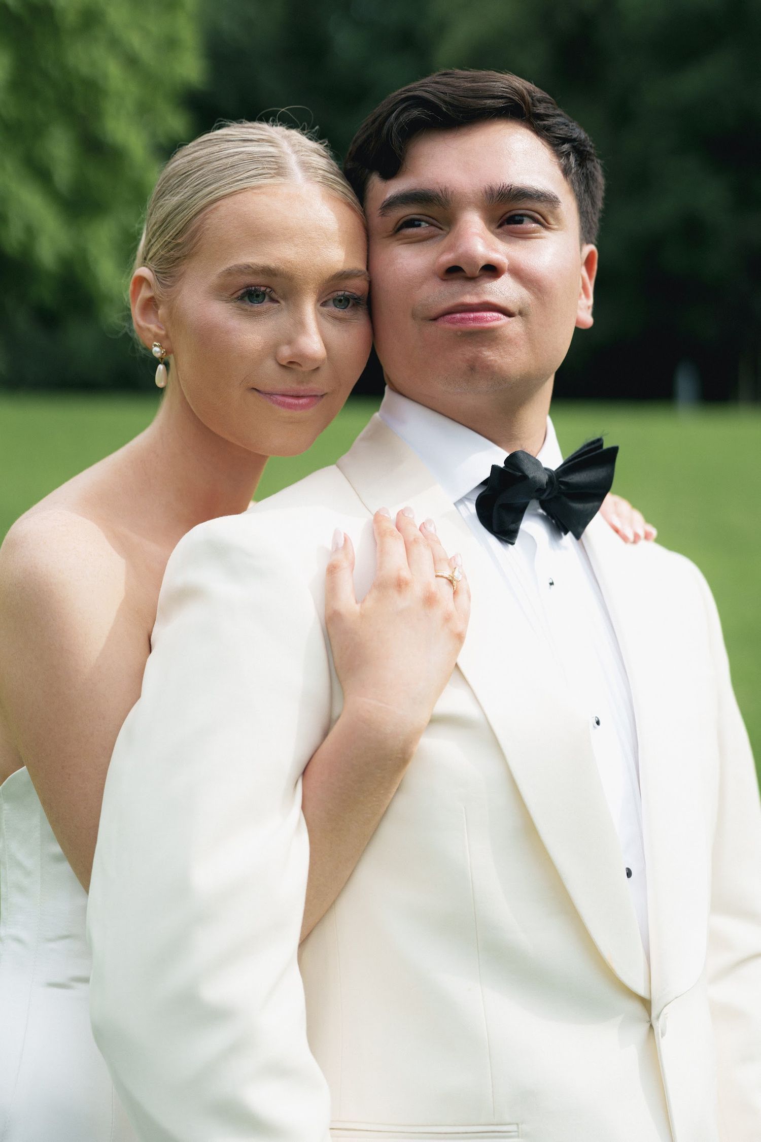 Smiling blonde bride in strapless gown leans on groom in white tuxedo with black bow tie outdoors.