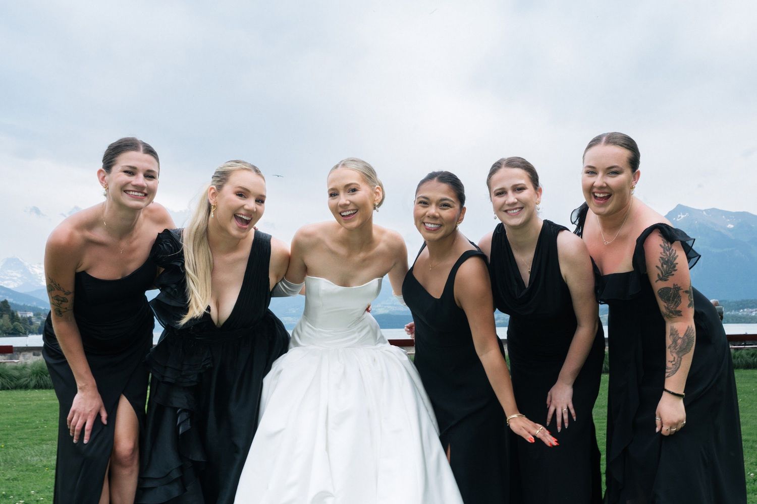 Bride laughing with five bridesmaids in black gowns on a lawn with mountain backdrop.