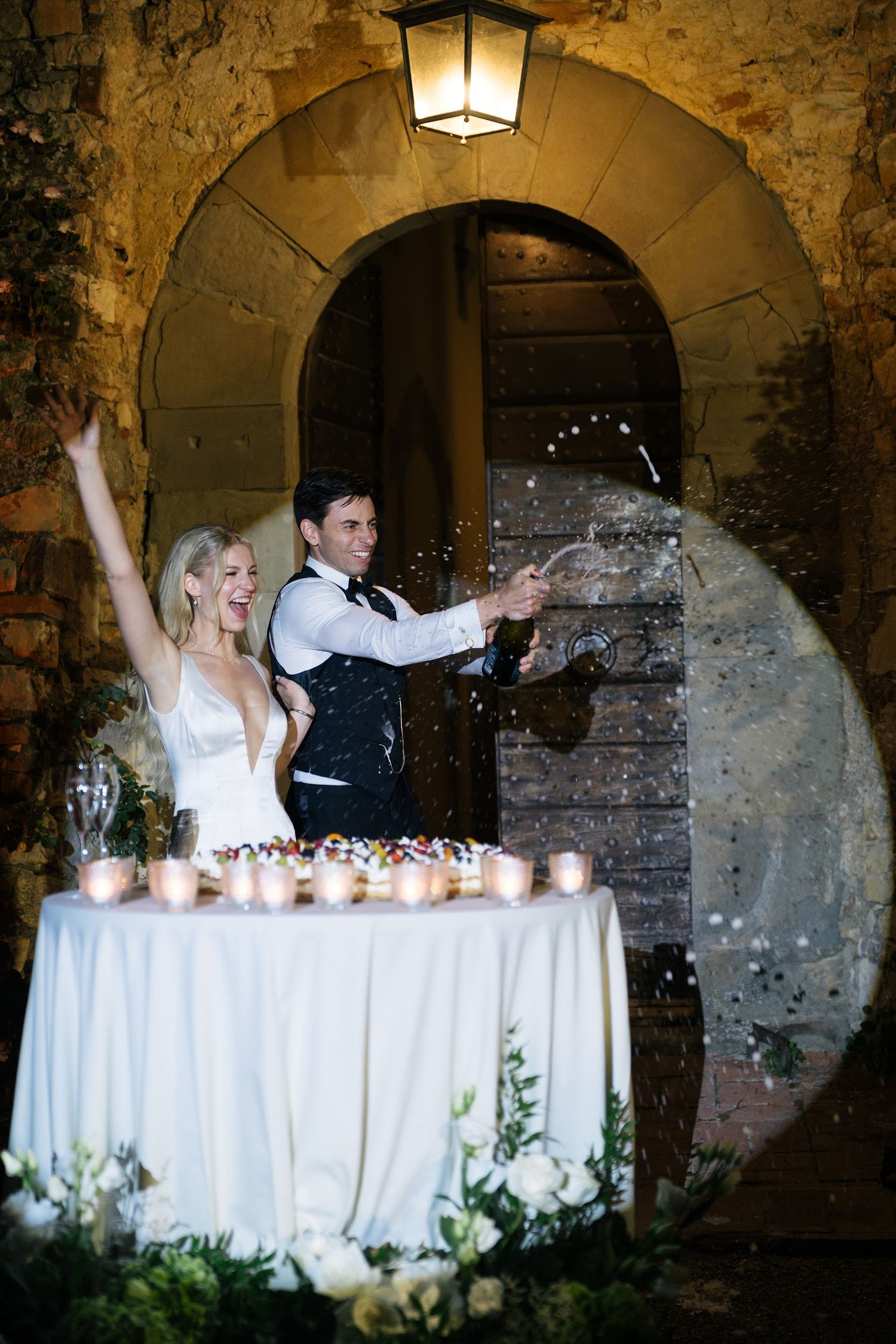 Bride and groom celebrating with champagne spray at candlelit Italian castle wedding reception.