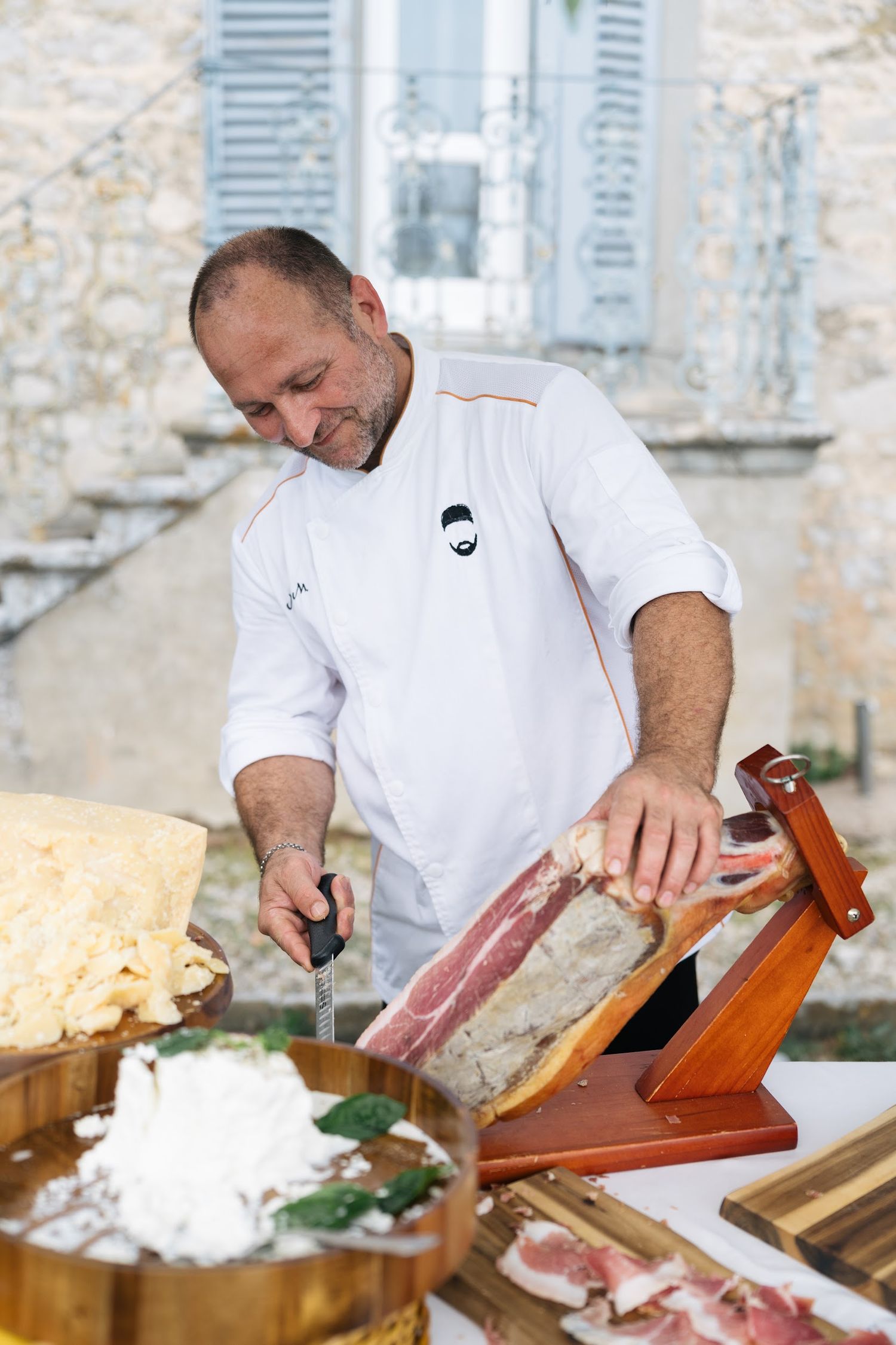 Chef in white coat slices prosciutto on a wooden stand at an elegant outdoor Italian wedding reception.
