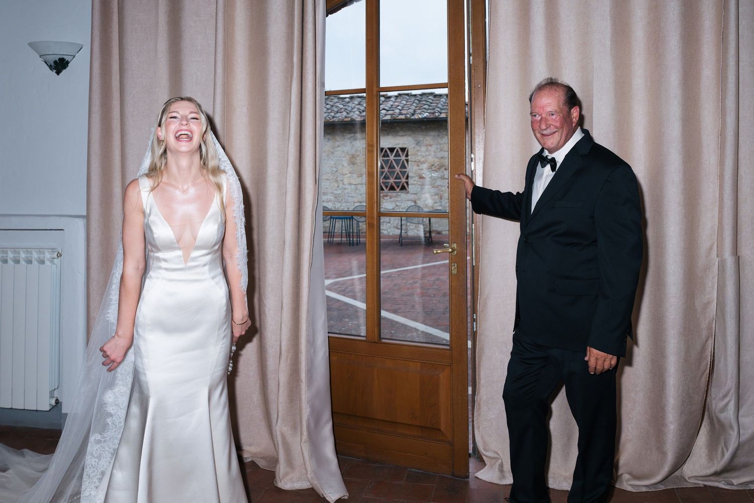 Bride in a satin gown laughs joyfully as an older gentleman in a tuxedo opens a door for her.