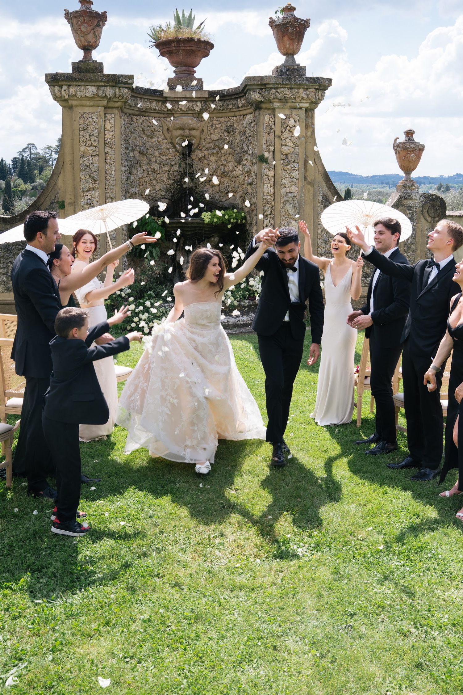 Wedding party celebrates newlyweds with flower petal toss on sunny lawn at elegant Italian villa.
