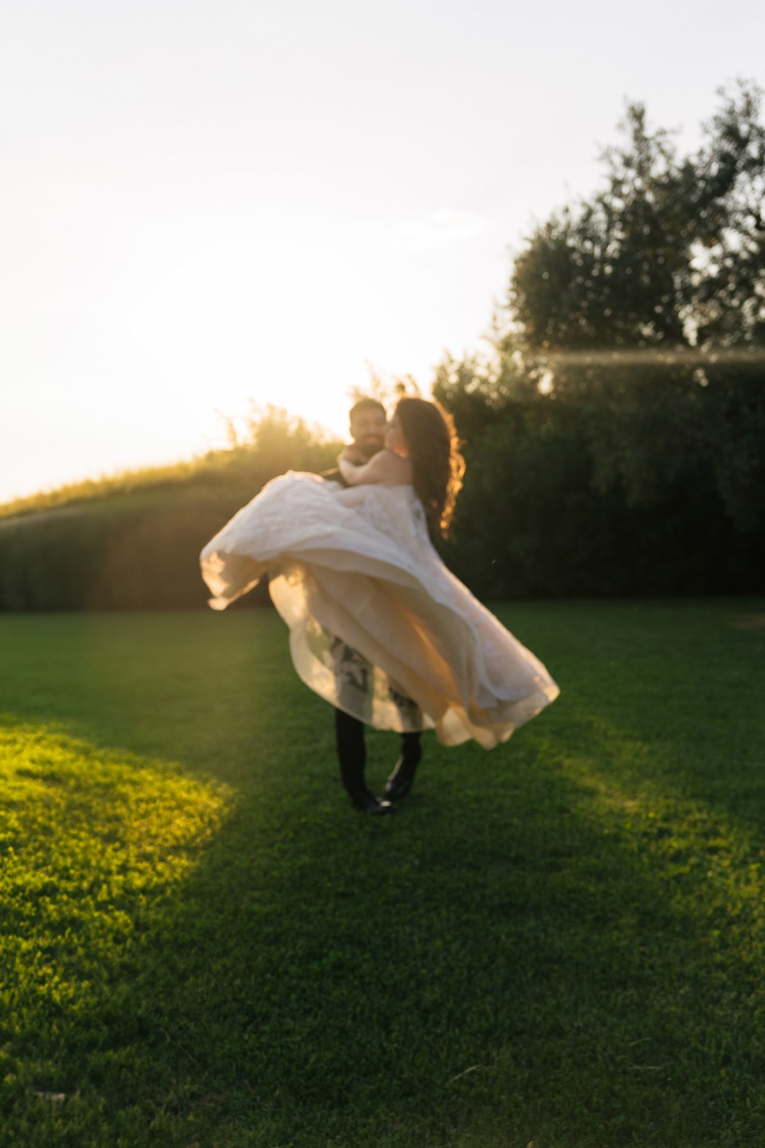 Groom lifting bride in flowing white dress at golden sunset on lush green lawn.