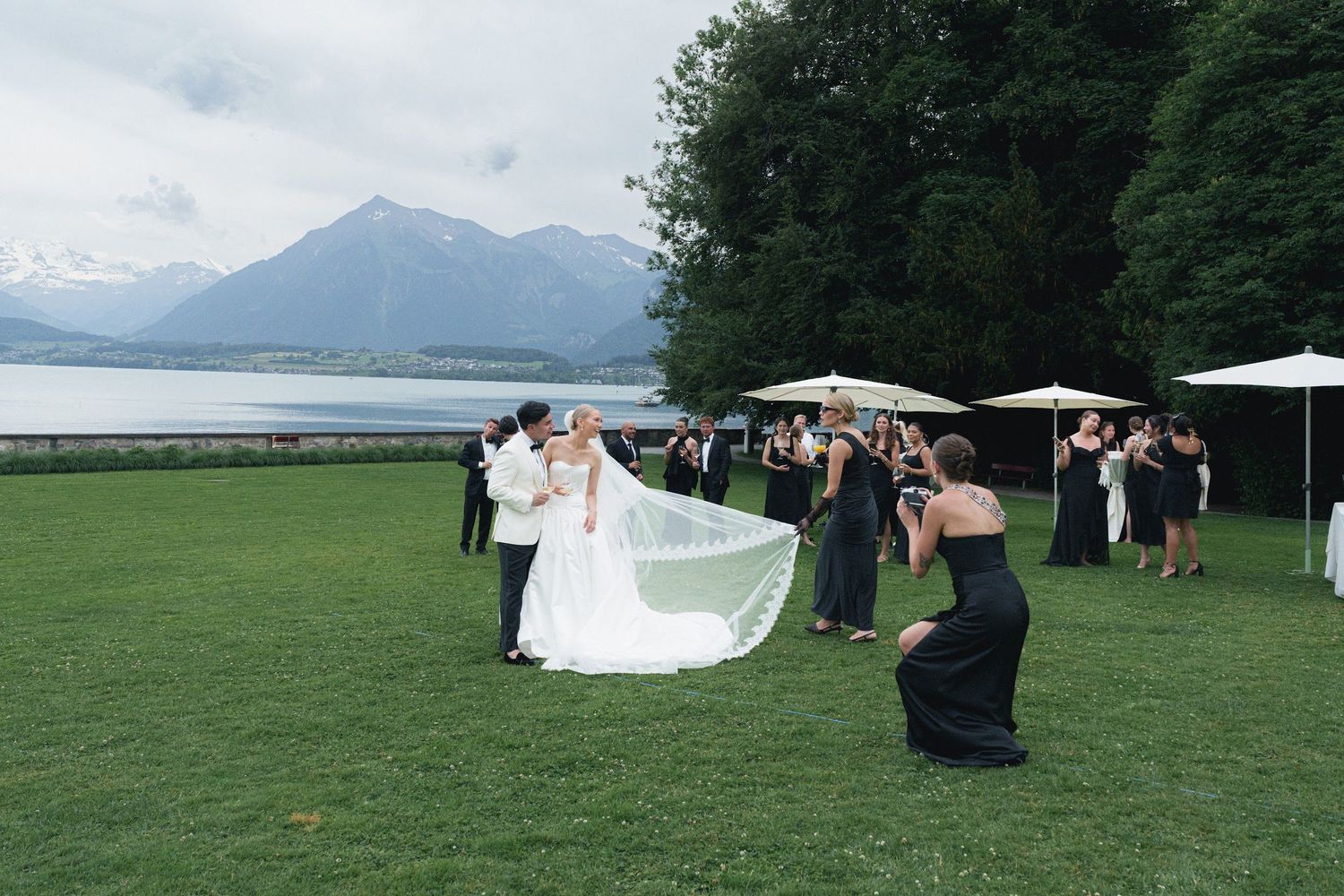 Wedding couple kissing on lawn as bridesmaid photographs them with mountains and lake behind.