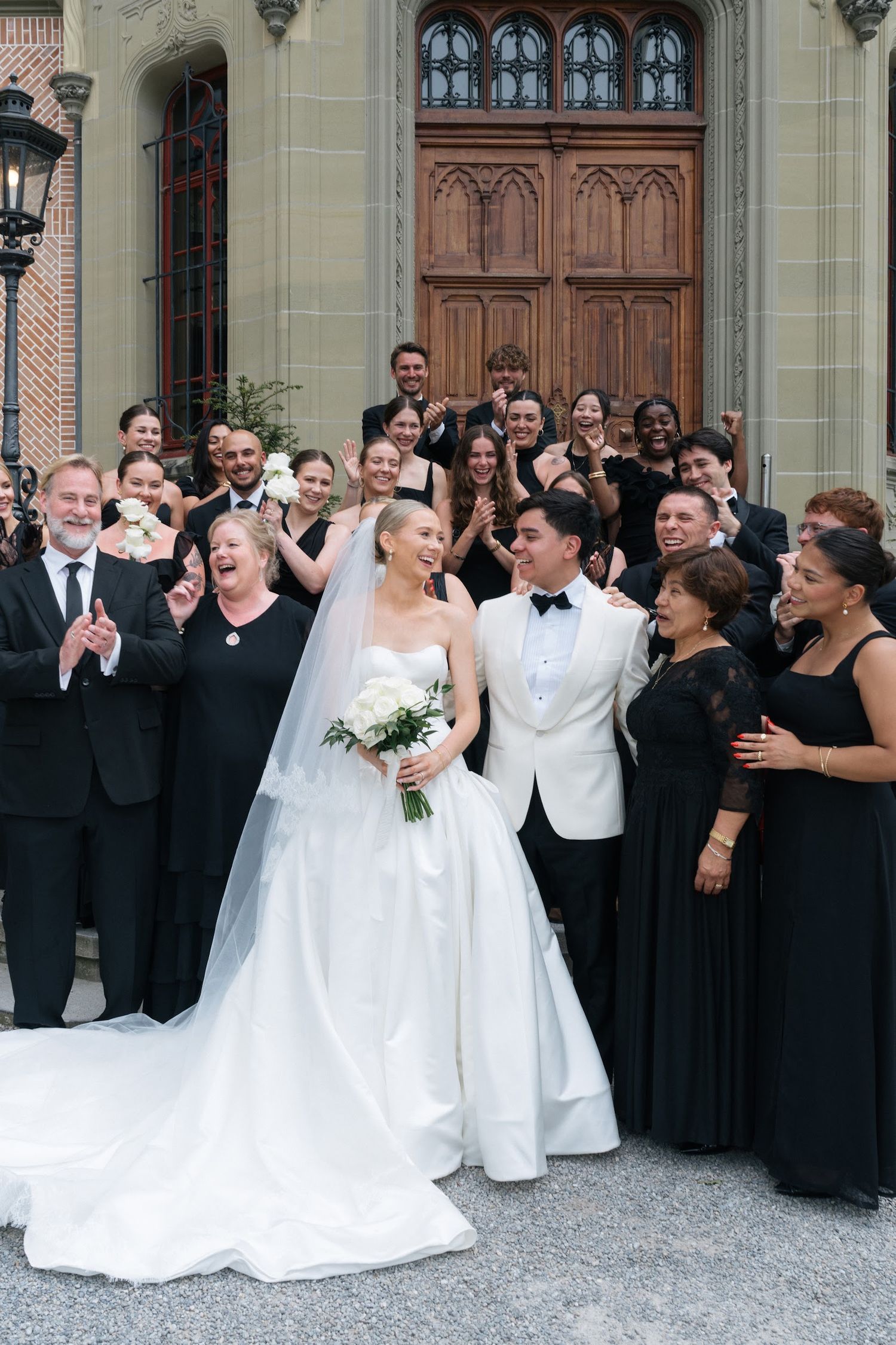 Joyful newlyweds celebrate with wedding party outside ornate wooden church doors, bride holding white bouquet in veil.